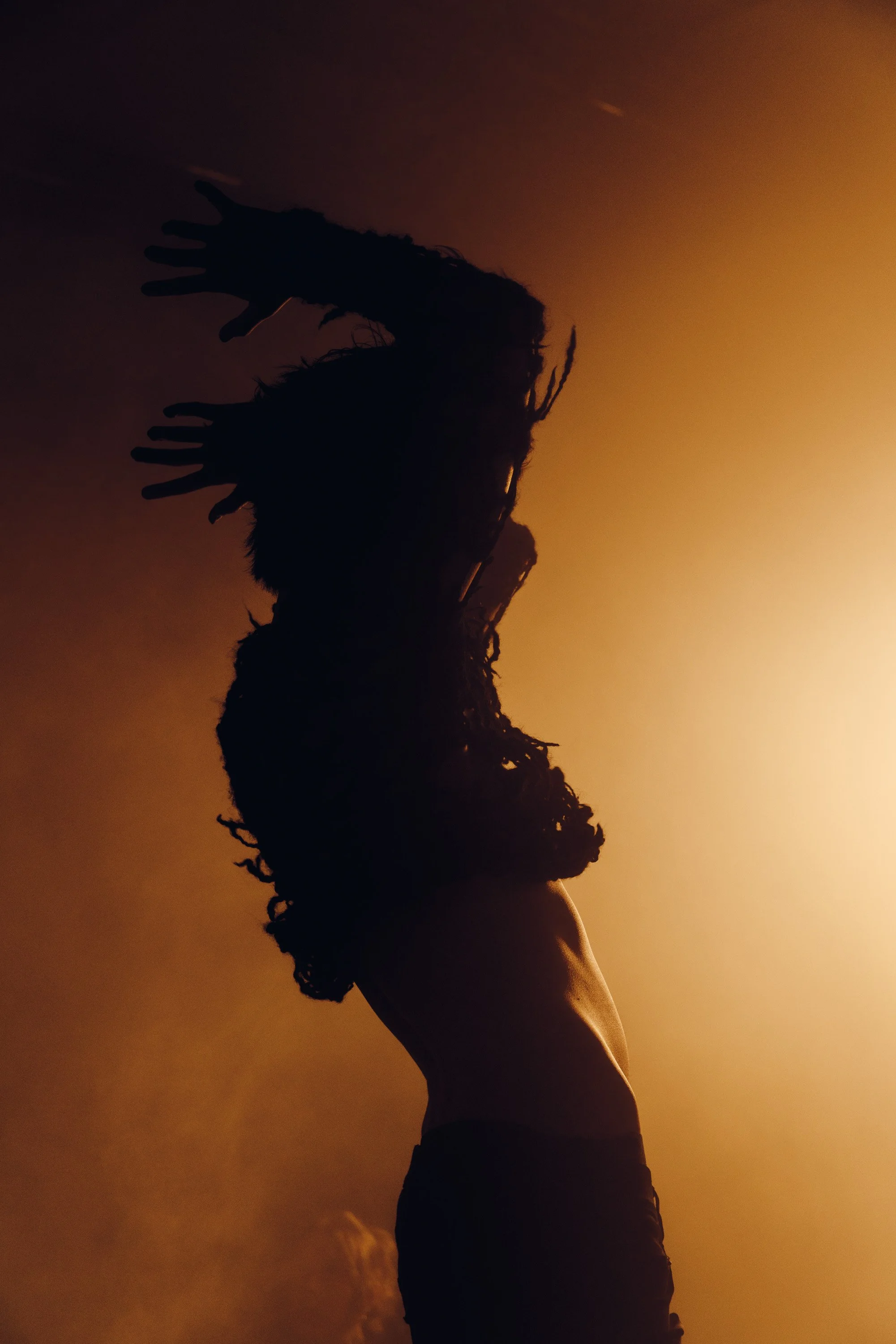 Silhouette of a person with dreadlocks standing against a warm, orange sunset sky.