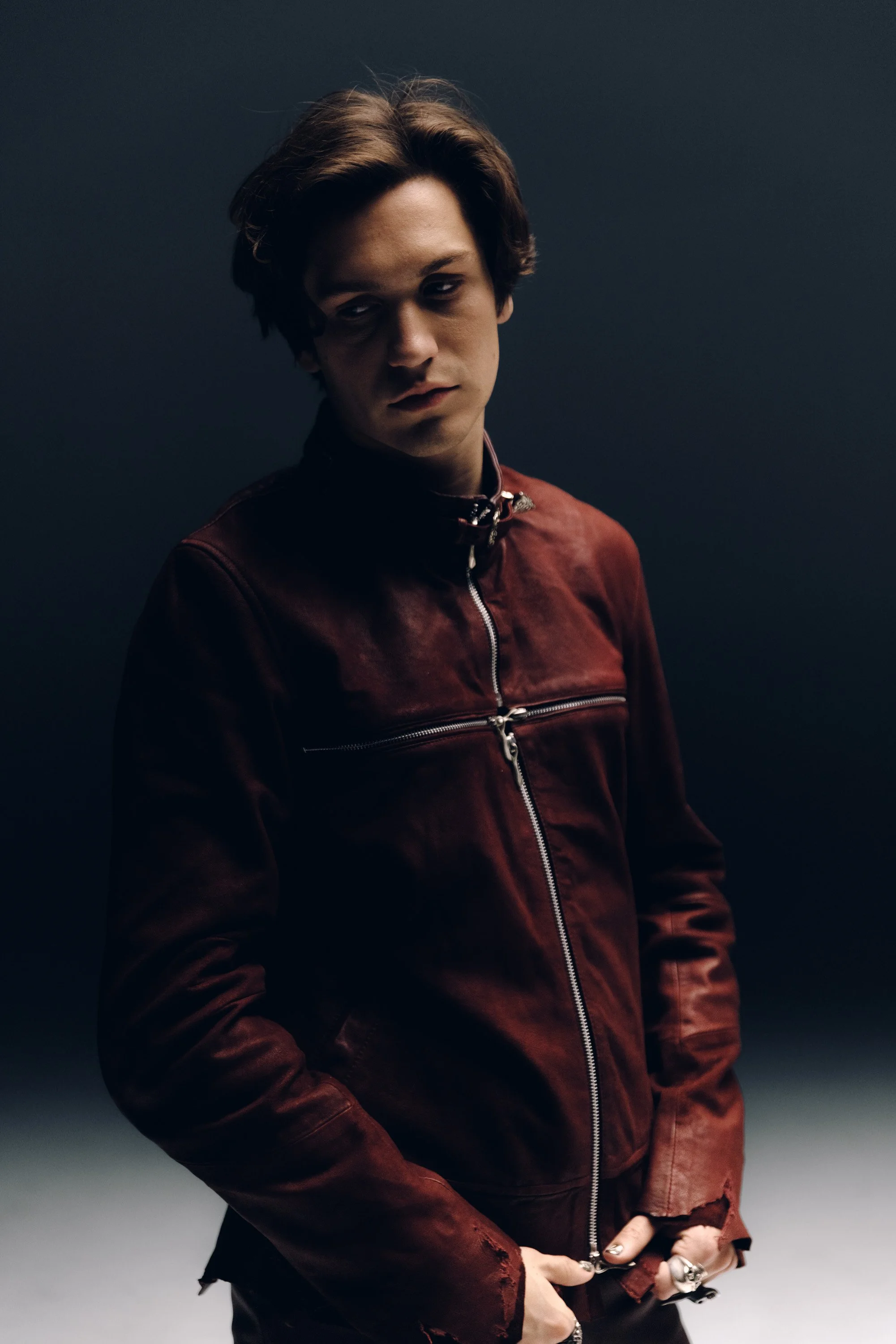 Young man with dark hair and fair skin wearing a red leather jacket with silver zippers, standing against a dark background.