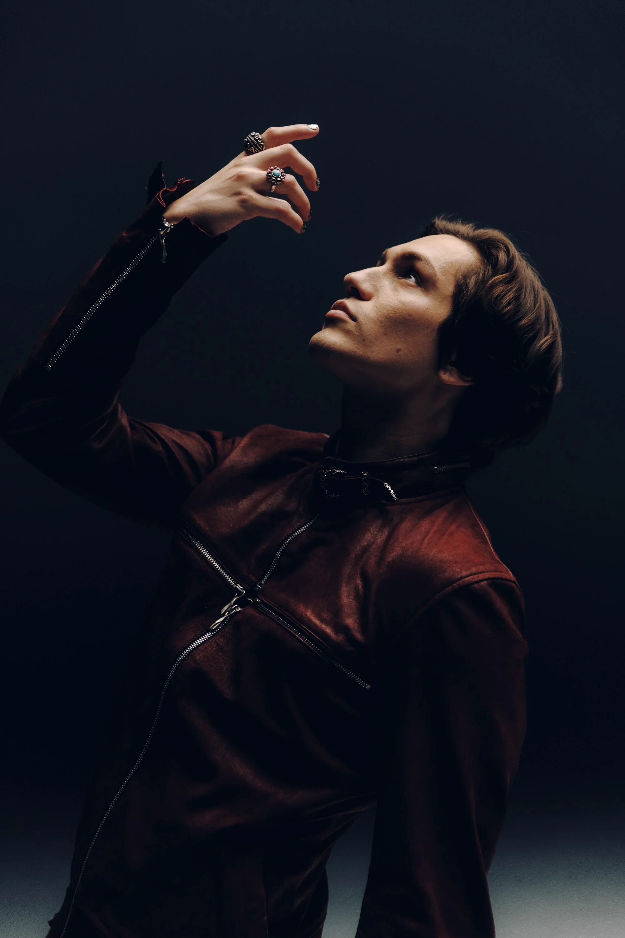A man with brown hair is wearing a brown leather jacket with zippers, looking up, with his right hand raised and wearing multiple rings, against a dark background.