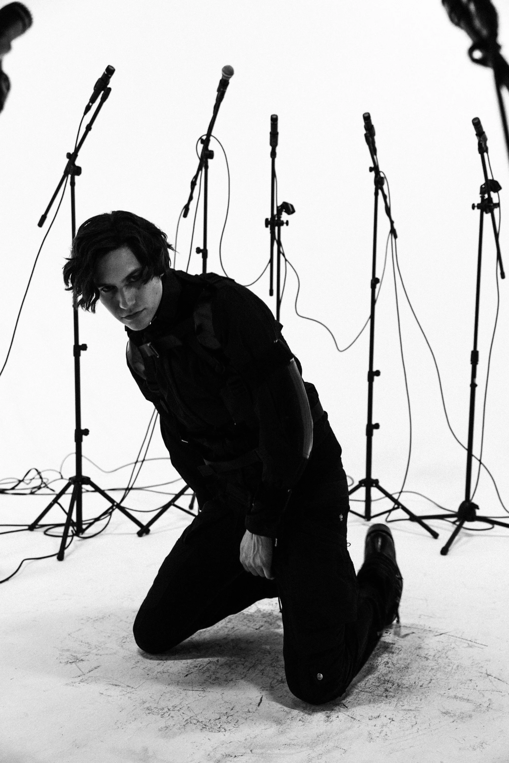 Black and white photo of a person with dark, wavy hair, kneeling on one knee on the floor in front of multiple microphone stands. The person is looking directly at the camera with an intense expression, wearing dark clothing. The background is plain 