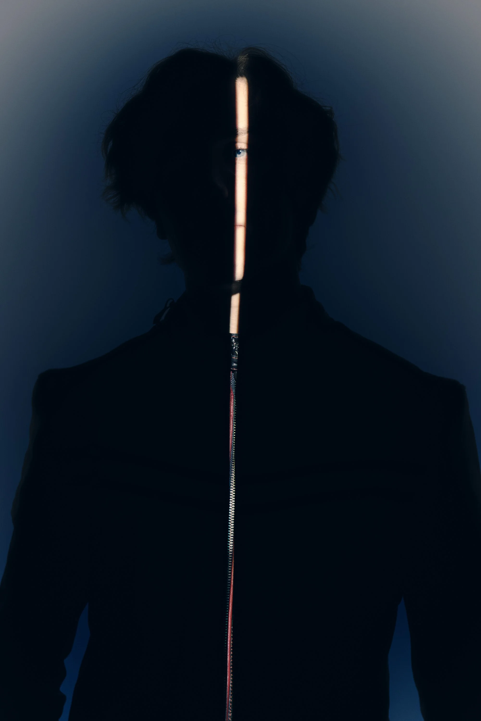A person wearing a black jacket with a zipper, standing in a dark environment. Light creates a vertical stripe across their face and torso, revealing only one eye and some facial features.