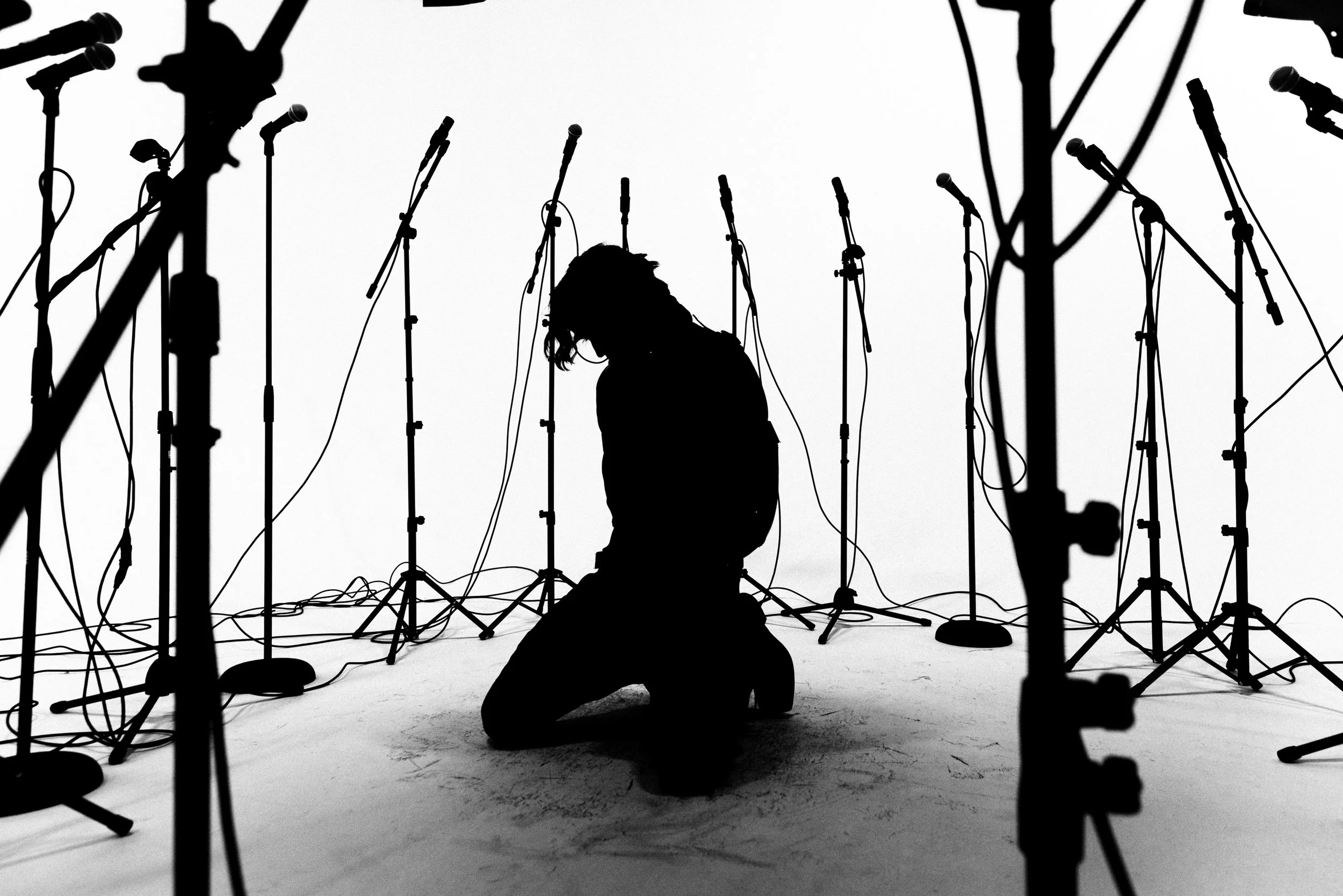 Silhouette of a person kneeling among multiple microphone stands in a studio with a white background.