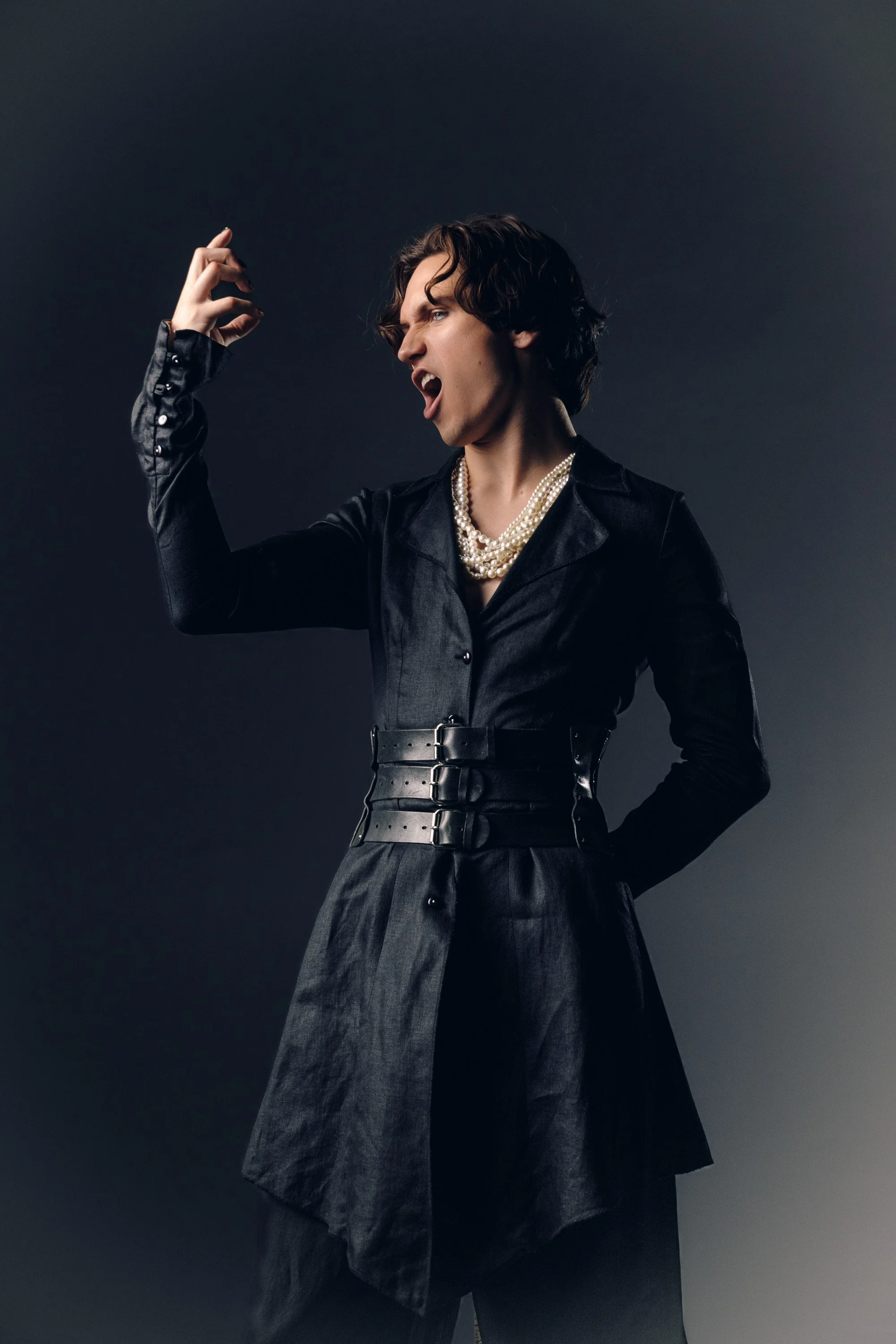 A woman with short curly hair wearing a black leather suit and pearl necklaces, posing with an angry expression and clenched fist against a dark background.