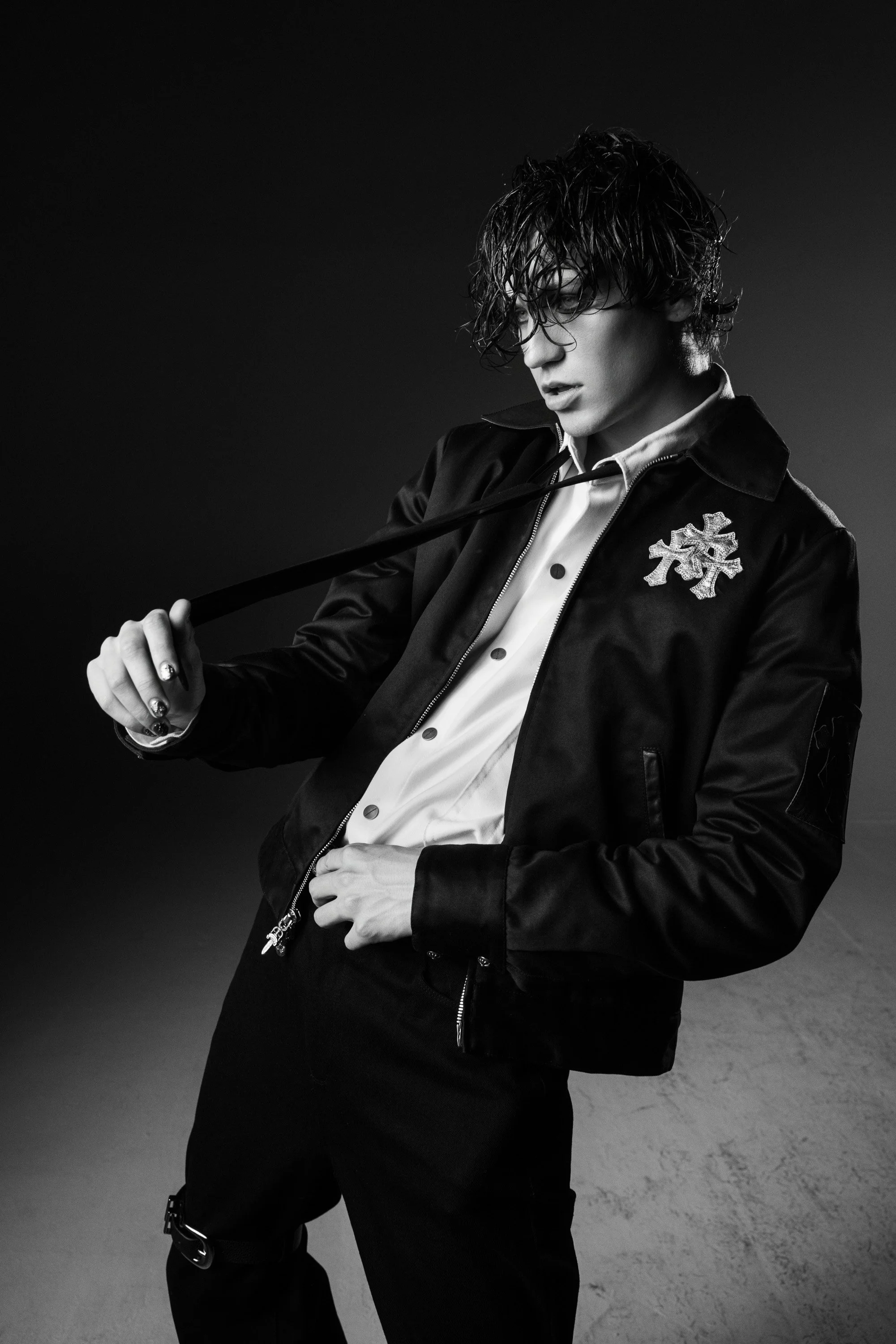 A young man with wet, curly hair and glasses wearing a black satin bomber jacket with a snowflake patch, white shirt, and black pants, holding a leash in a studio with a dark background.
