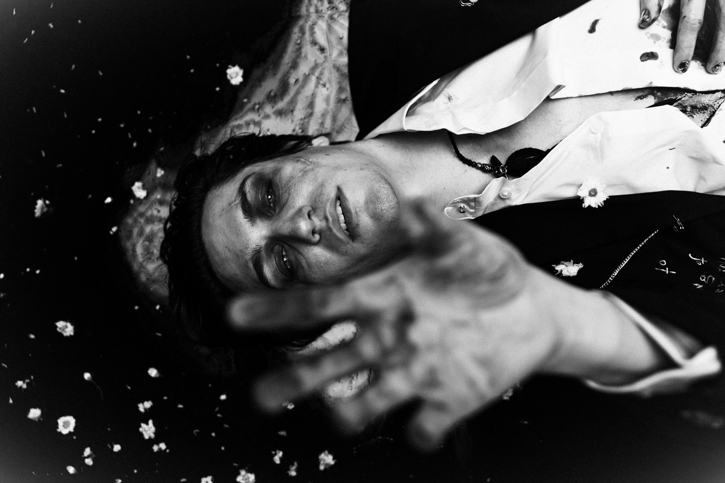 A woman lying on her back with a joyful expression, surrounded by scattered flower petals, wearing a white top and a black necklace, with her hand resting on her chest.