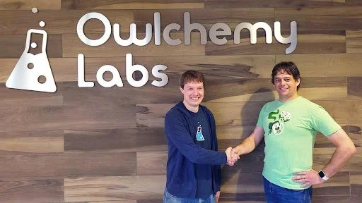 Two men stand in front of a wooden wall with an “Owlchemy Labs” sign, smiling and shaking hands, suggesting a professional or celebratory moment in a studio or office setting.