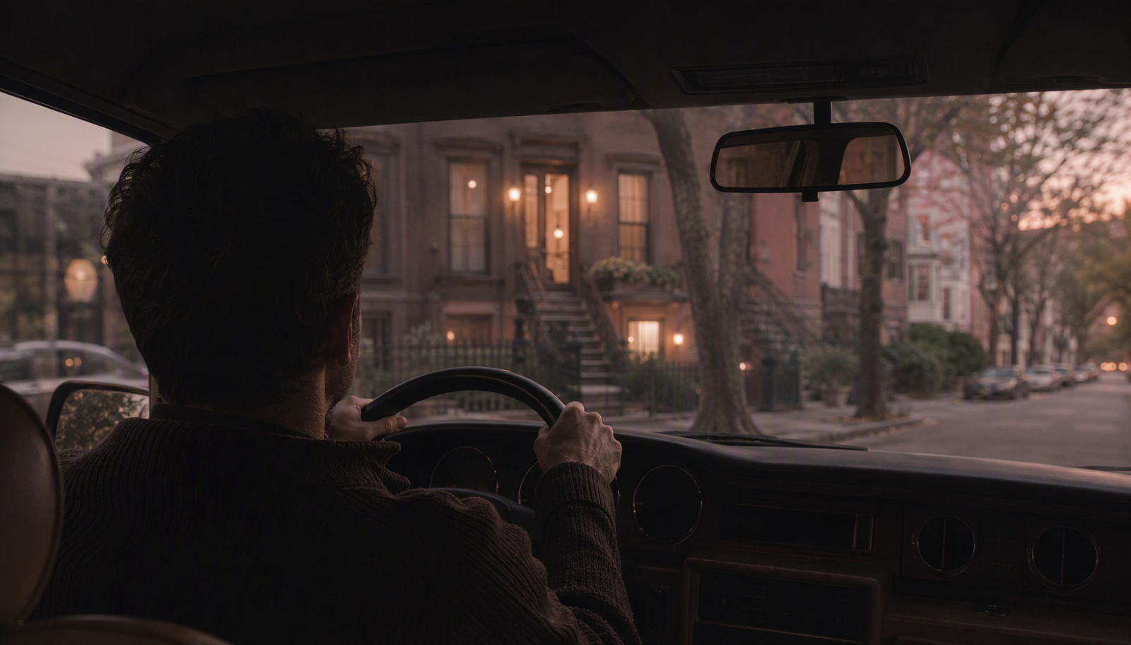 A man sitting in his parked car on a Brooklyn brownstone street at dusk, hands on the wheel, taking a few minutes before going inside