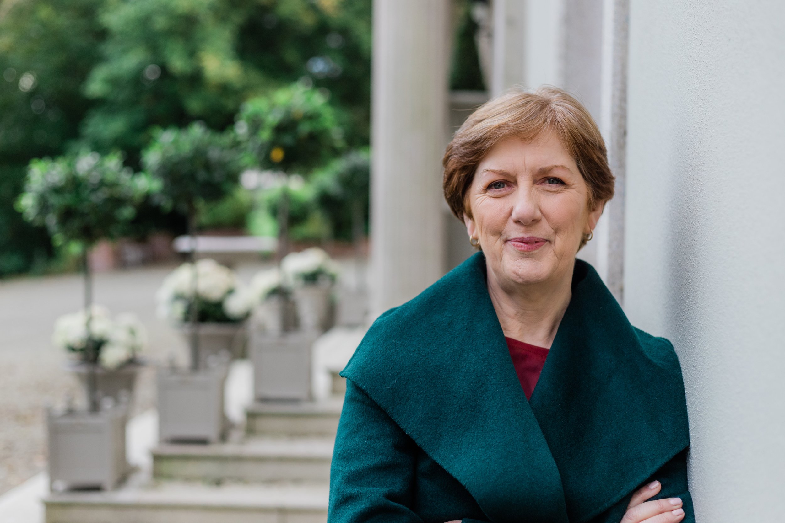 Mary Pyne with short brown hair smiling outdoors, wearing a teal coat, standing near a white wall with potted plants and greenery in the background.
