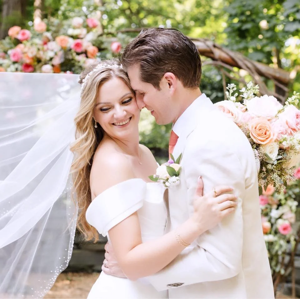 A young woman with shoulder-length auburn hair and blue eyes stands outdoors near a body of water with blurred hills in the background.