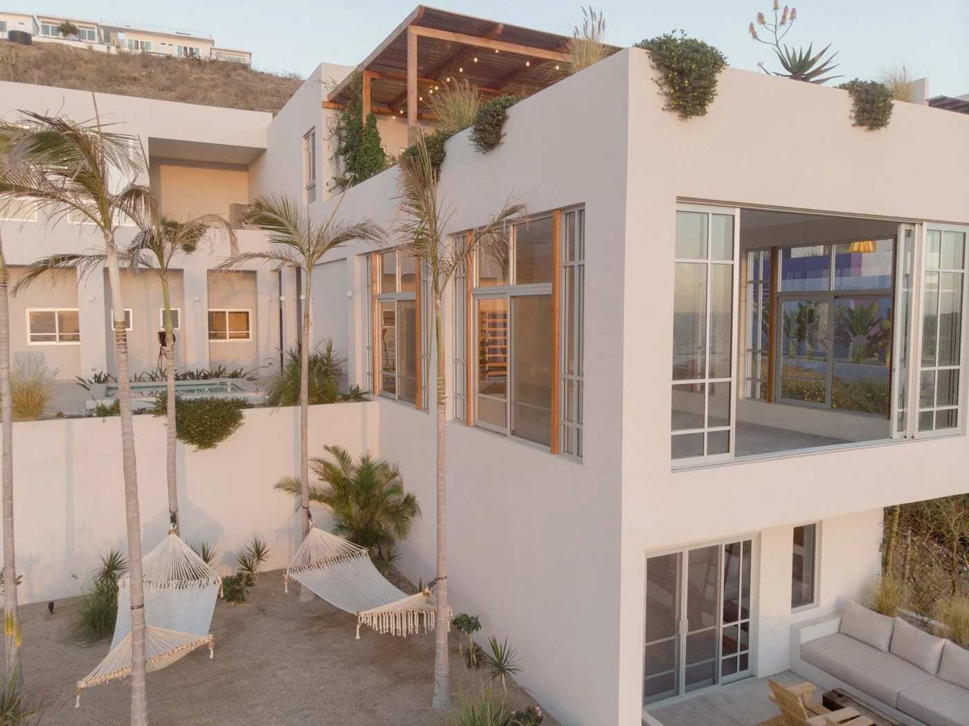 Modern white multi-story house with large glass windows, surrounded by palm trees, outdoor hammocks, and patio furniture, at sunset.