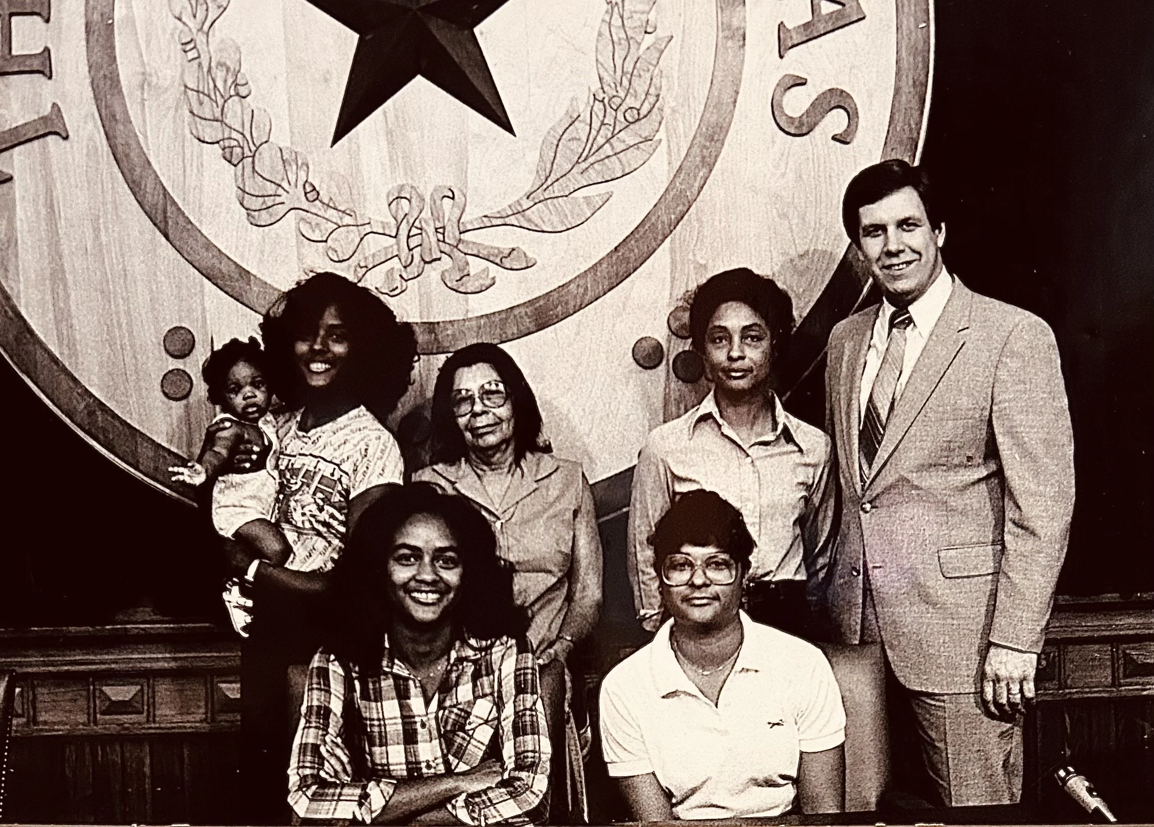 Black and white photo of Faith, including four women, Faith's mother, grandmother, and sisters, posing indoors in front of a large emblem of the seal of the Texas State Bar.
