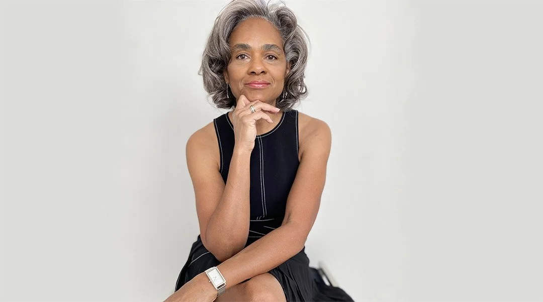 Faith James wearing a sleeveless black dress, sitting against a plain white background, looking at the camera with a thoughtful expression.