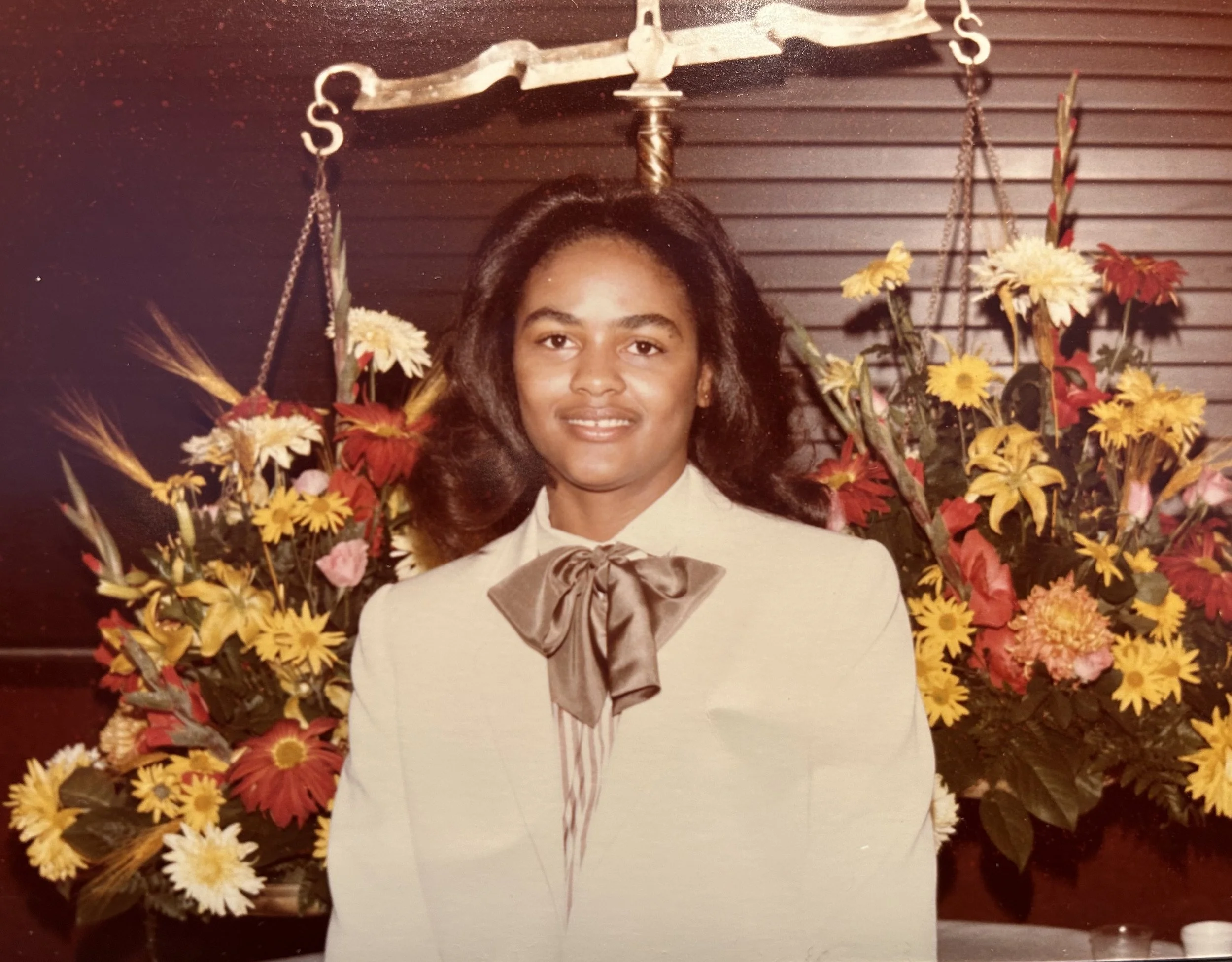 Faith is standing in front of a dark wooden backdrop decorated with yellow, pink, and red flowers. She is wearing a light-colored blazer with a large bow at the collar.