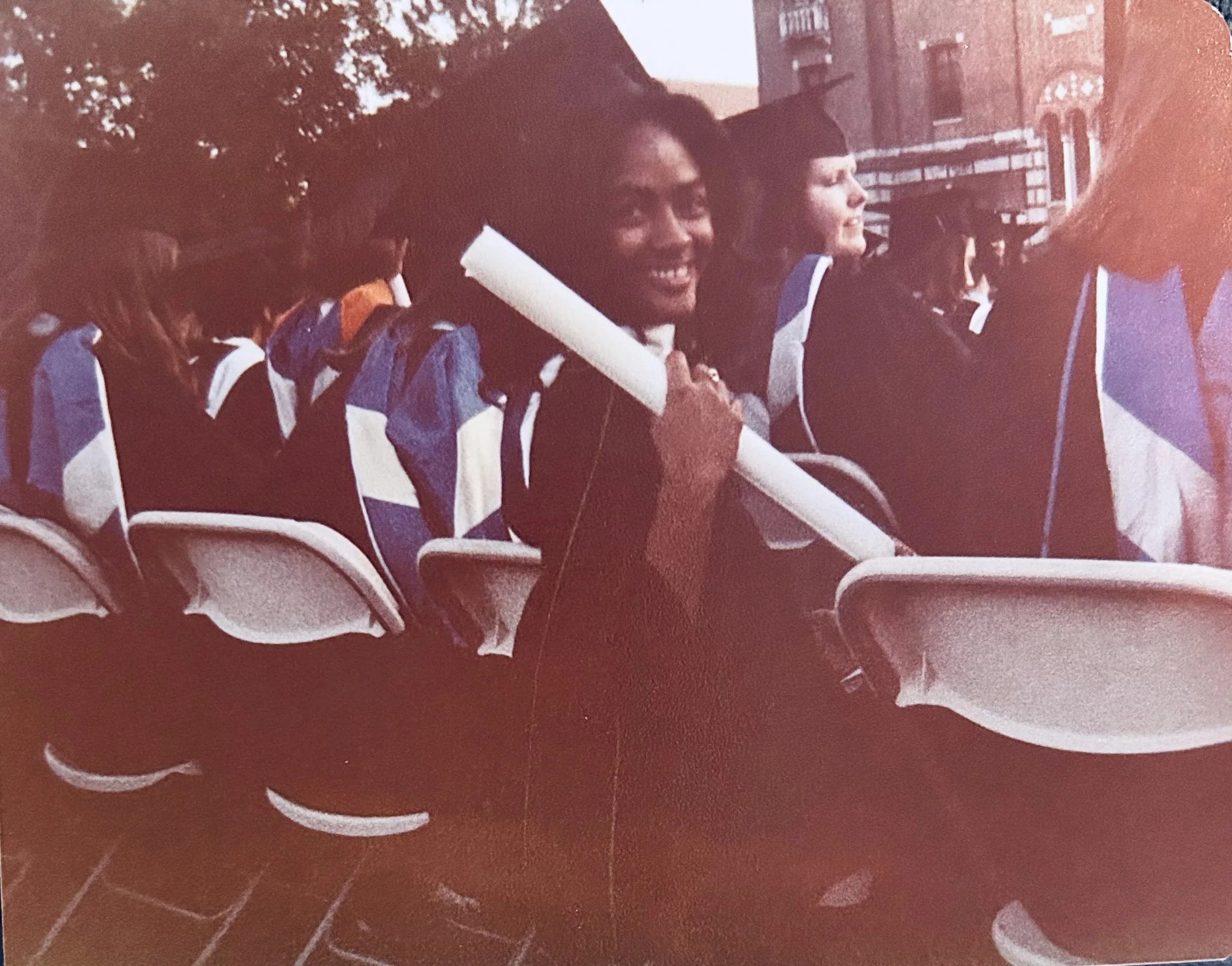 Group of young women in graduation gowns sitting outdoors, Faith James holding a rolled-up diploma and smiling.