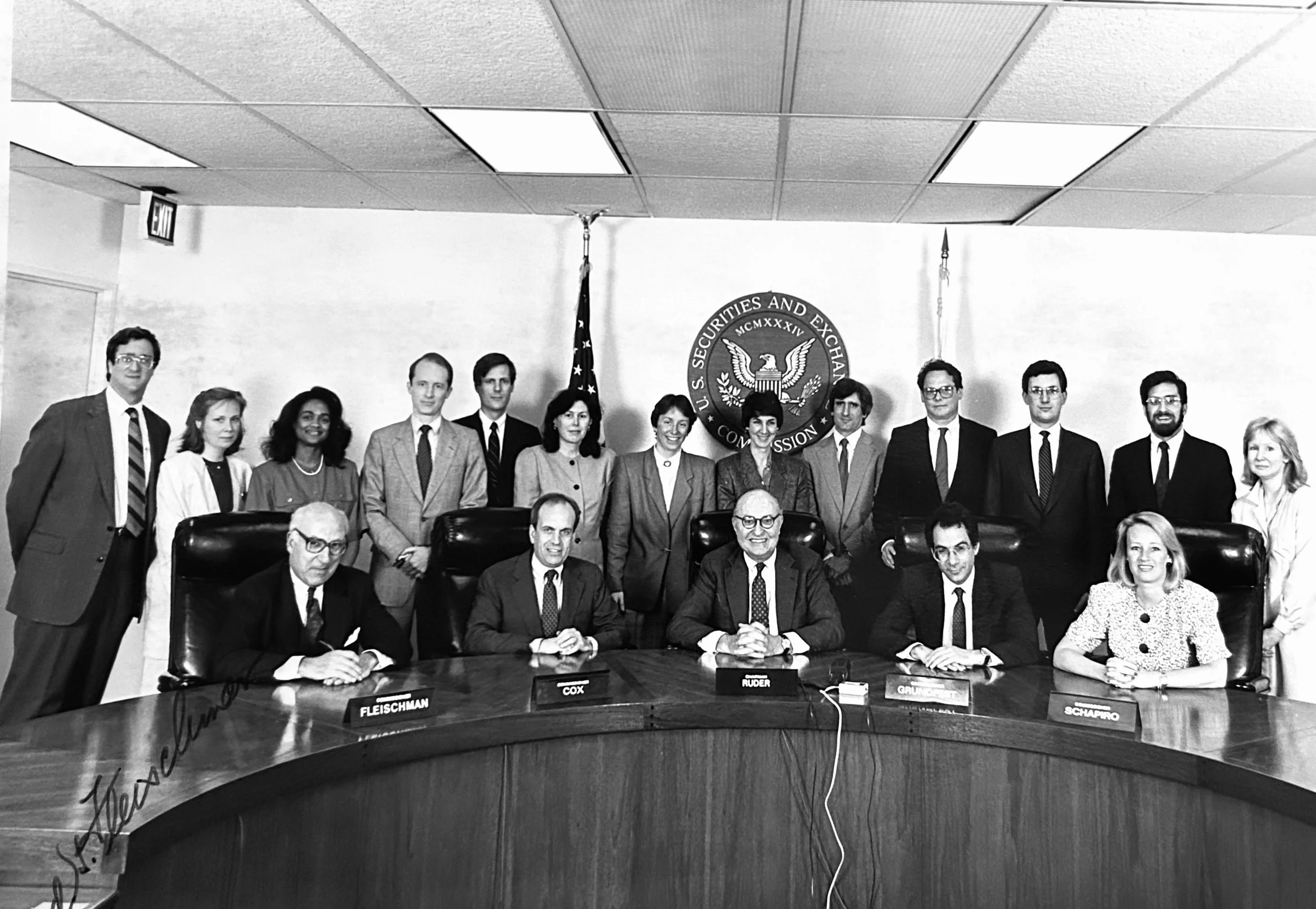 Black and white group photo of 15 people in business attire, seated and standing behind a conference table, in front of the U.S. Securities and Exchange Commission emblem, with American flags on either side.