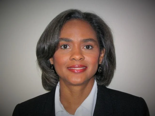 Portrait of Faith James with shoulder-length gray hair, wearing a black blazer and white shirt, smiling against a plain background.