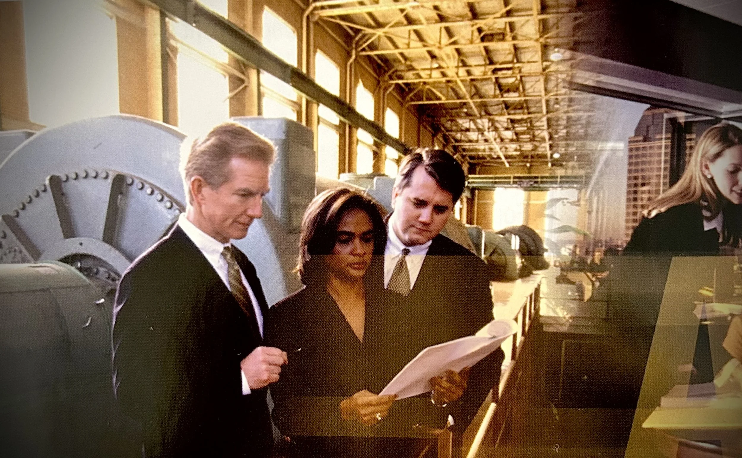 Three professionals, two men and one woman, in formal attire, reading documents in an office with large windows and exposed ceiling beams.