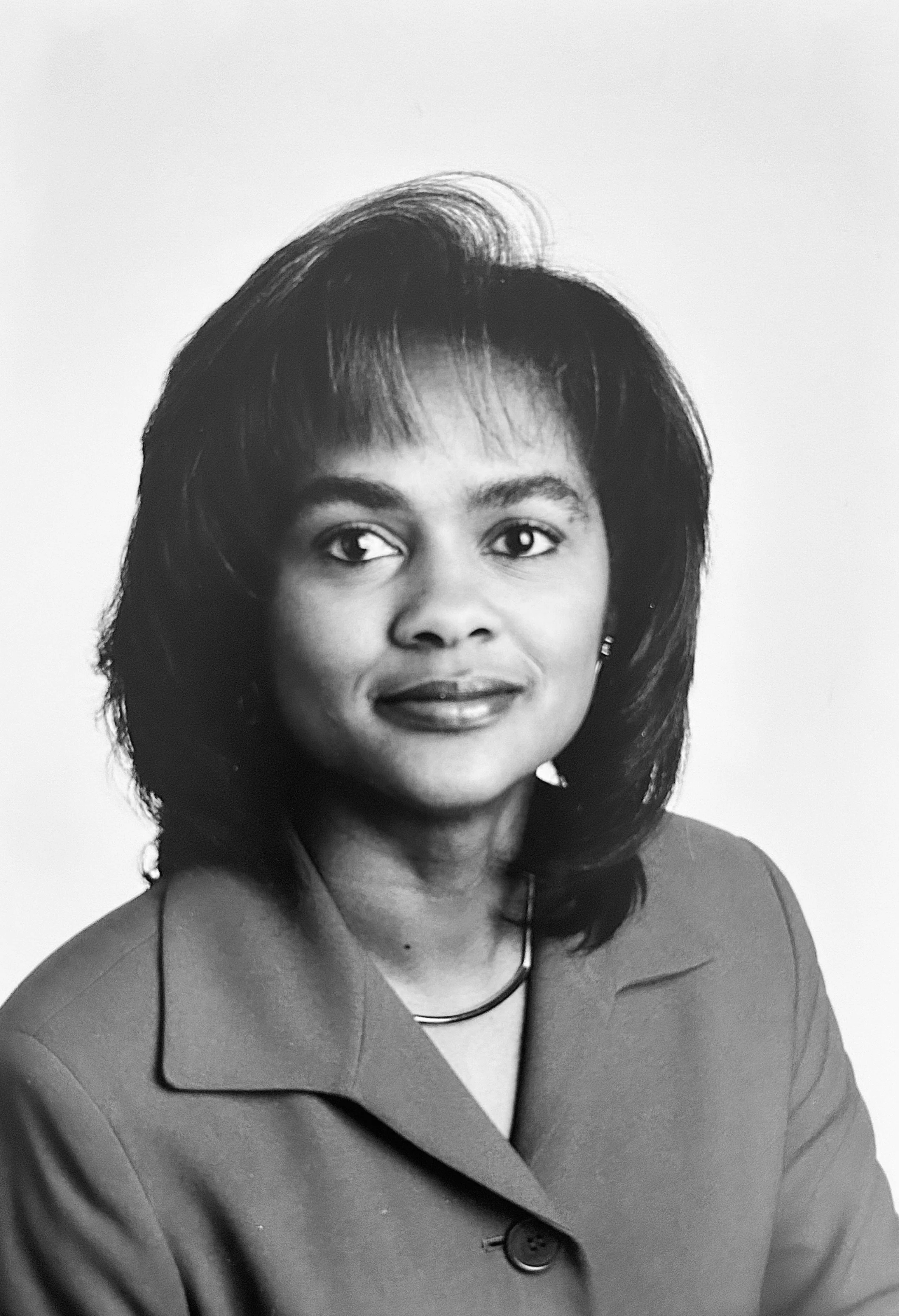 Black and white portrait of a woman of Faith with shoulder-length hair wearing a blazer and a necklace. She is looking slightly to the right with a neutral expression.