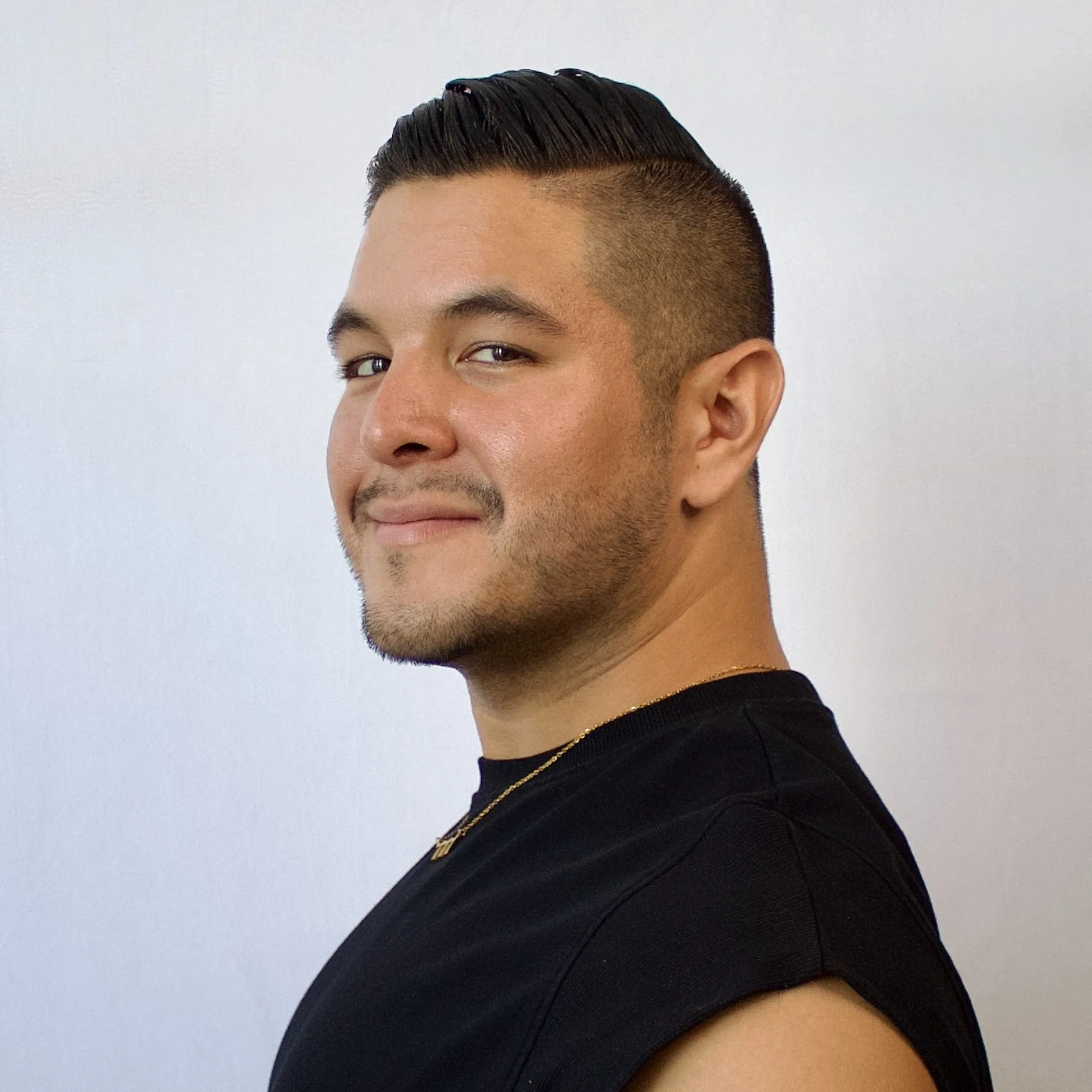 A young man with short, slicked-back dark hair and a slight beard, wearing a black shirt and a gold chain necklace, standing against a plain white background.