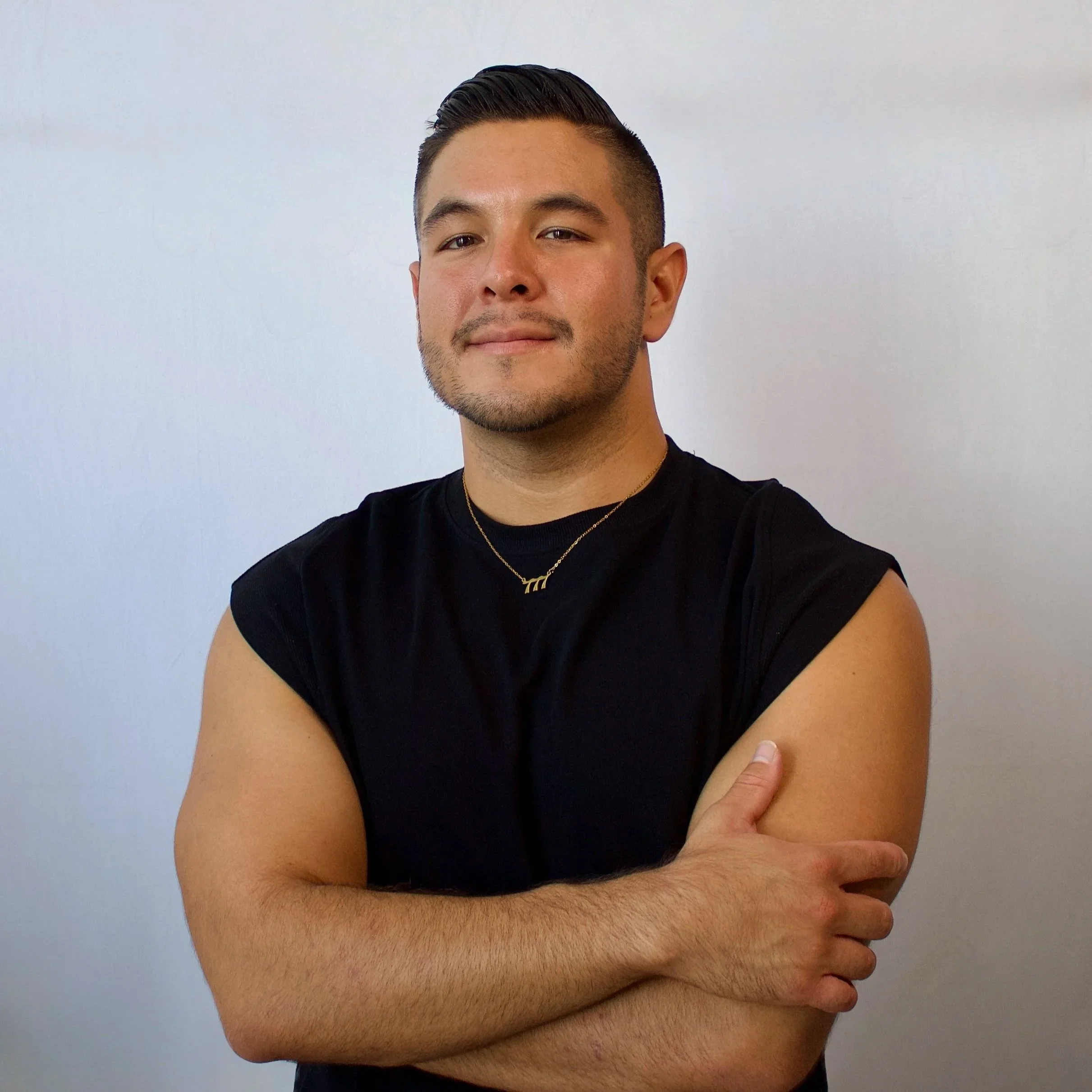 A young man with short dark hair, styled upwards, and facial hair, is smiling slightly. He is wearing a black sleeveless shirt and gold necklaces. His arms are crossed in front of his body, and he is standing against a plain white wall background.