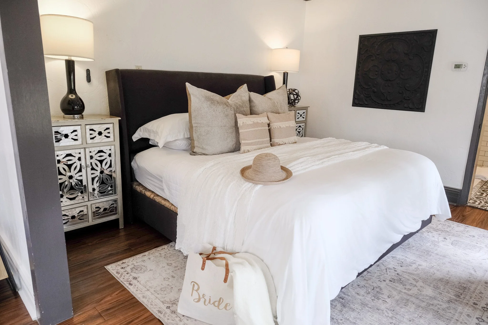 The Bliss Suite bedroom features white bedding, decorative pillows, and a straw hat on the bed. A mirrored nightstand with a lamp is beside the bed, and a "Bride" bag sits on the floor. Wall art is visible.