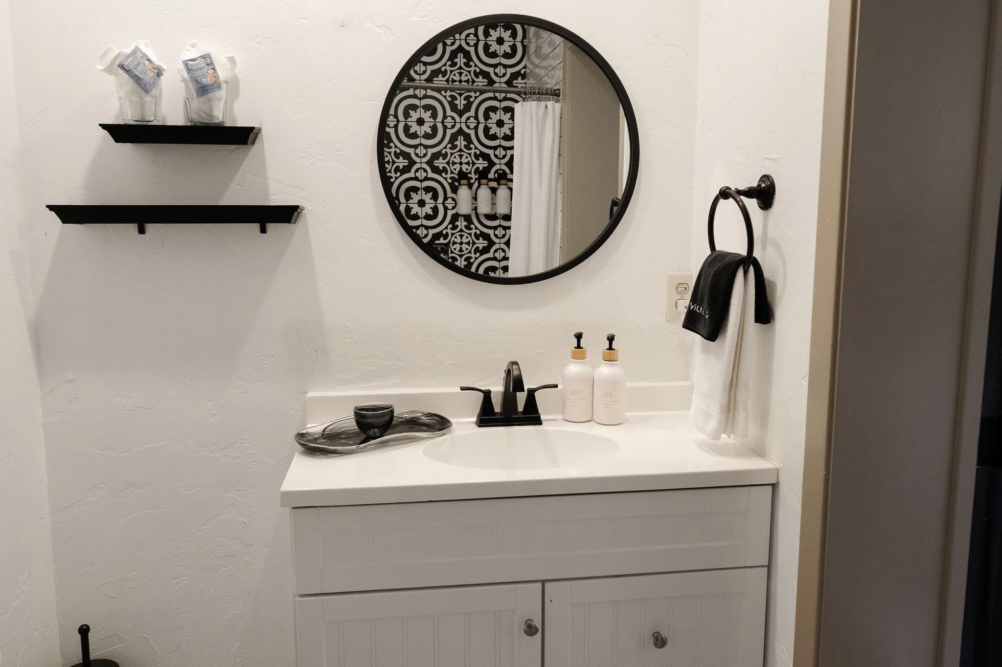 Modern bathroom with a white vanity, black faucet, and round mirror. Two decorative soap dispensers, a black towel on a ring, and two shelves with wrapped glasses are visible. Tiled shower and curtain are reflected in the mirror.
