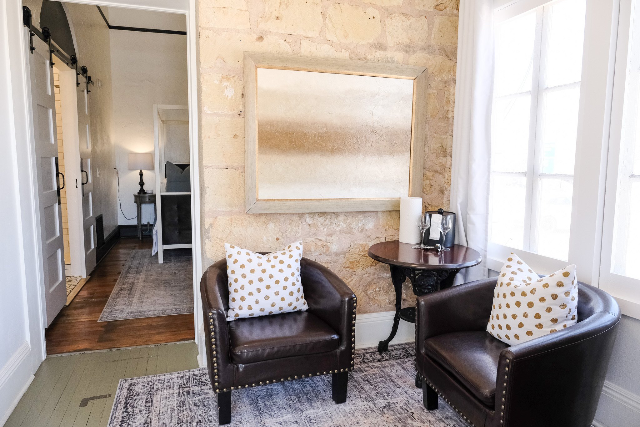 Cozy sitting area with two brown leather chairs, polka dot cushions, small round table with wine essentials, large framed art on stone wall, and doorway leading to a bedroom.