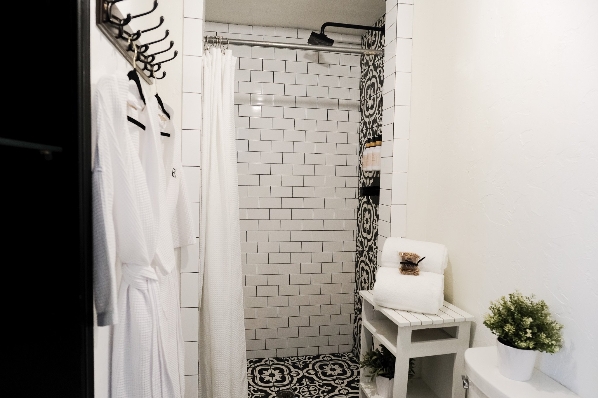 Modern bathroom with subway tiles, black and white patterned floor, white shower curtain, two plush white robes, and a small table with folded towels and plants.