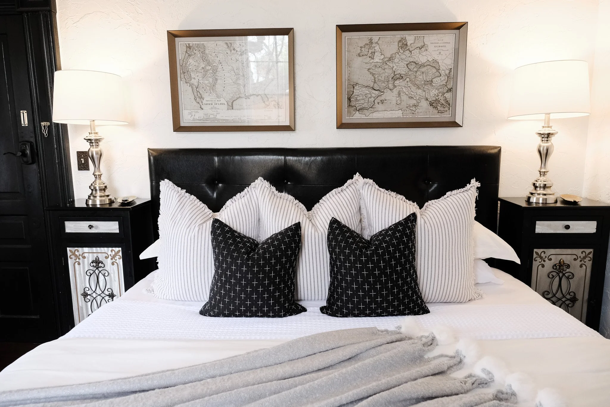 The Bella Suite bed featuring multiple pillows in black and white, flanked by two nightstands with silver lamps, and two framed maps on the wall above the headboard.