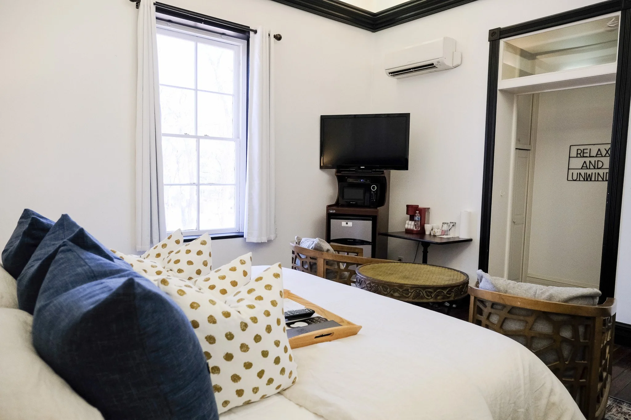 A cozy bedroom with a bed featuring polka dot pillows, a window with white curtains, a wall-mounted TV, and an air conditioning unit. There is a small table with an in room coffee maker and cups, as well as decorative chairs.