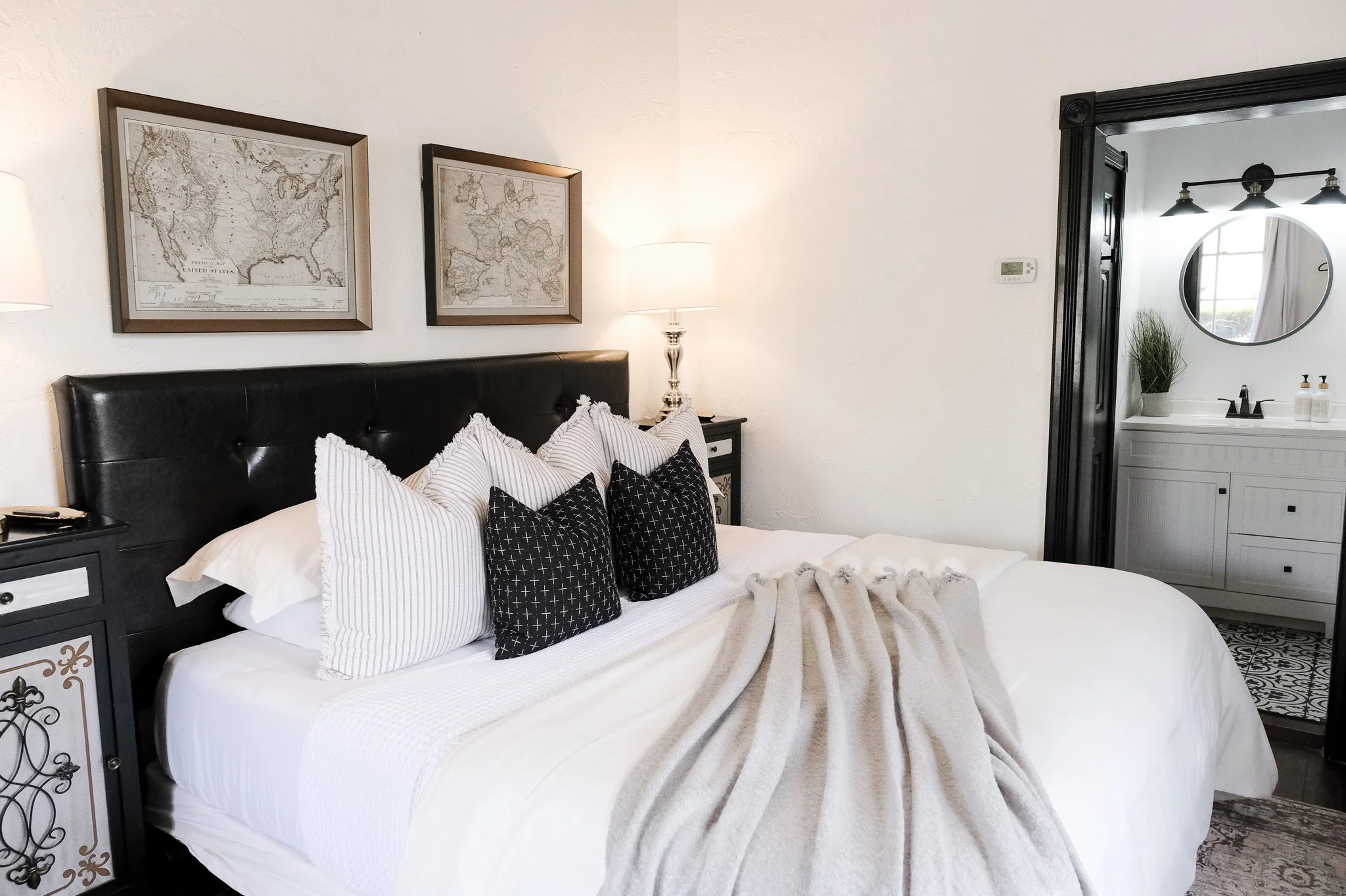 The Bella Suite features a plush bed, patterned pillows, two framed maps above the headboard, nightstand with a lamp, and view into a bathroom with a round mirror and sink.
