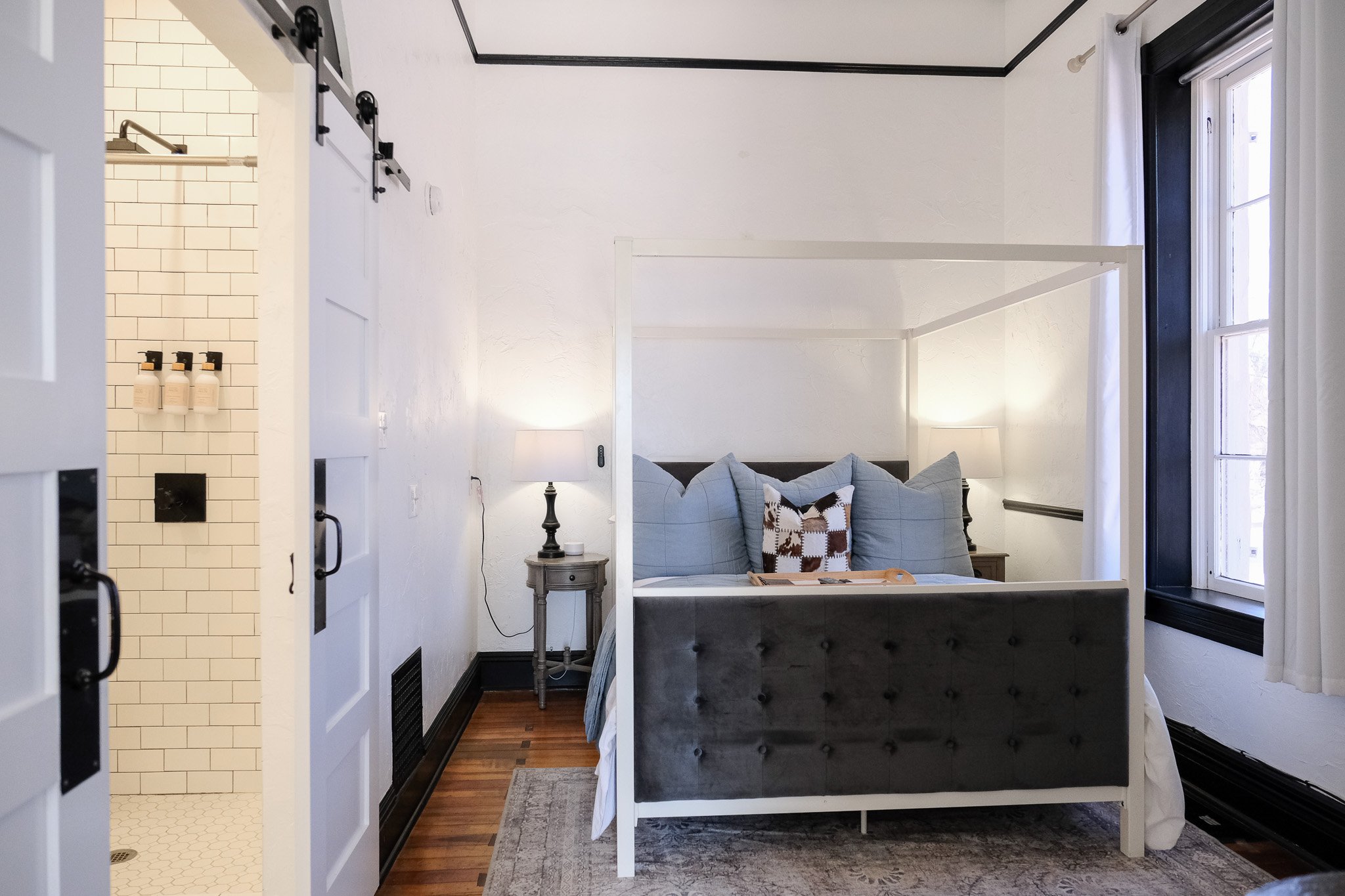 The Tuscan Suite at The Evers House has a modern bedroom with a canopy bed featuring blue accents, a side table with a lamp, and hardwood flooring. A sliding barn door reveals a bathroom with white subway tiles and a shower.