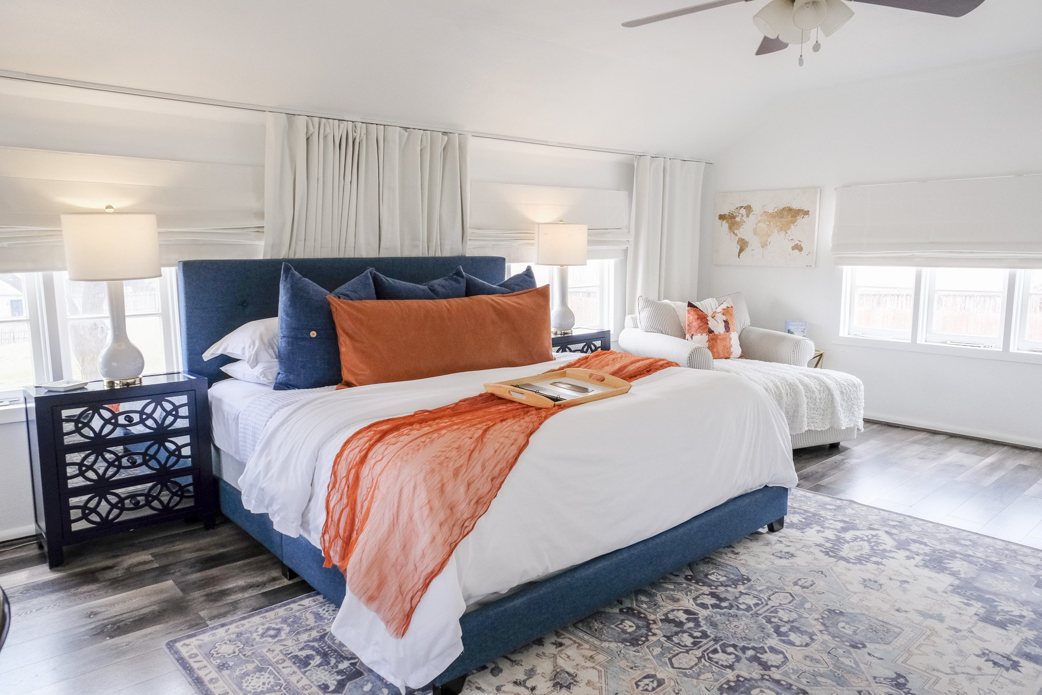 The Treehouse Suite features an upper level bedroom with a blue upholstered bed, side tables with lamps, and a white armchair. Light wood flooring and a patterned rug paired with large windows and a ceiling fan give it beautiful natural lighting.
