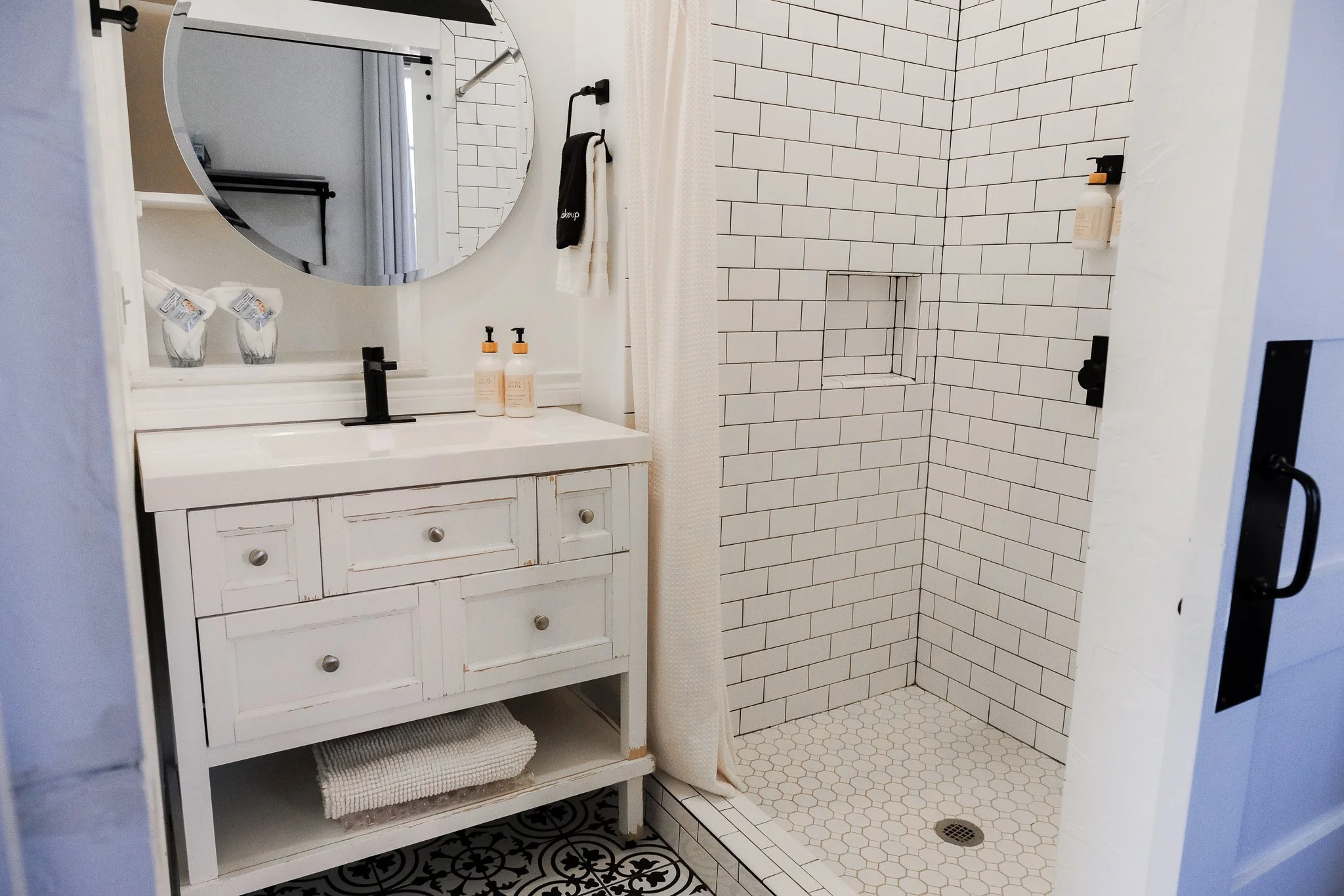 Seventh Heaven Suite bathroom at The Evers House featuring a white vanity with black faucet, round mirror, and patterned tile flooring. The walk-in shower includes white subway tiles, a built-in shelf, and folded towels with luxury bath products.