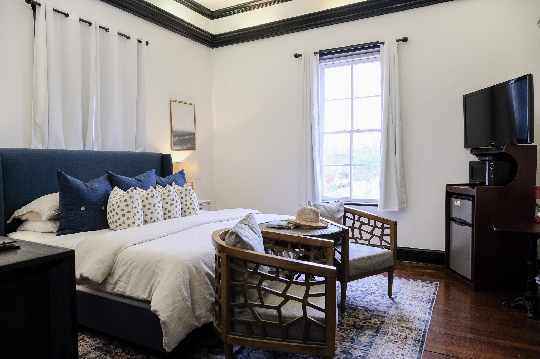 Cozy bedroom with a blue upholstered bed, multiple pillows, and white bedding. Two wooden chairs and a small table are in front of the bed. A window with white curtains is on the wall, and a TV with a microwave and mini-fridge is in the corner.
