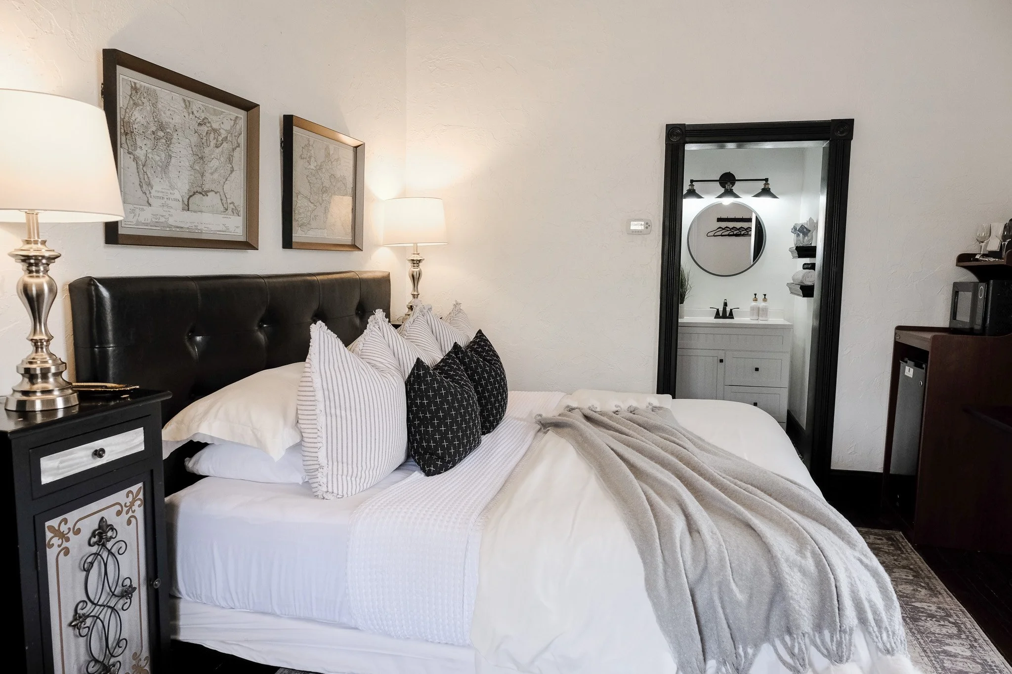 Cozy bedroom with king-sized bed, white and striped pillows, gray blanket, map artwork, nightstand with lamp, and view of bathroom with vanity and round mirror.