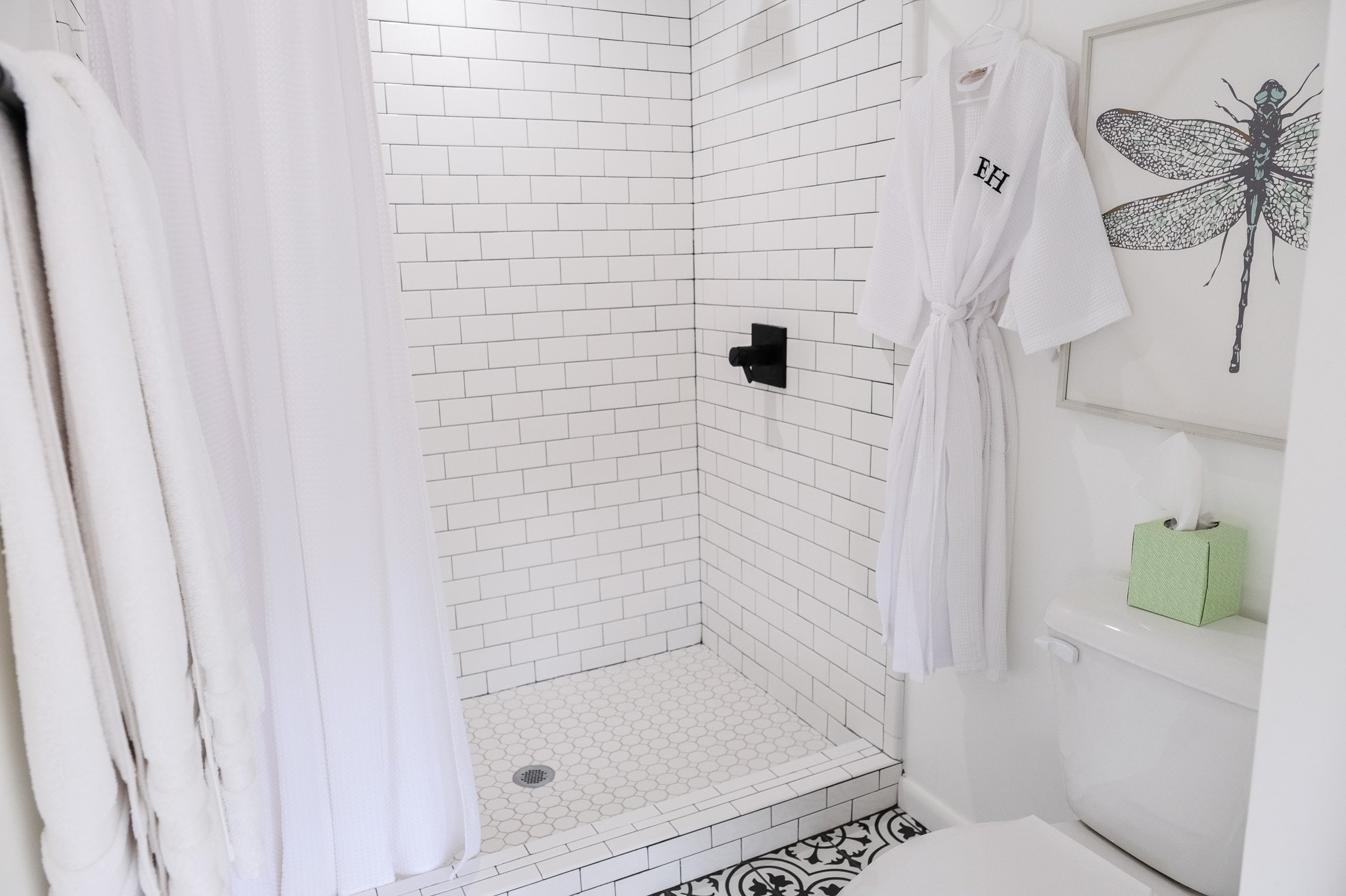 The Treehouse Suite features a modern bathroom with white subway tile shower, black shower fixtures, plush robe, framed dragonfly art, and a green tissue box on a toilet.