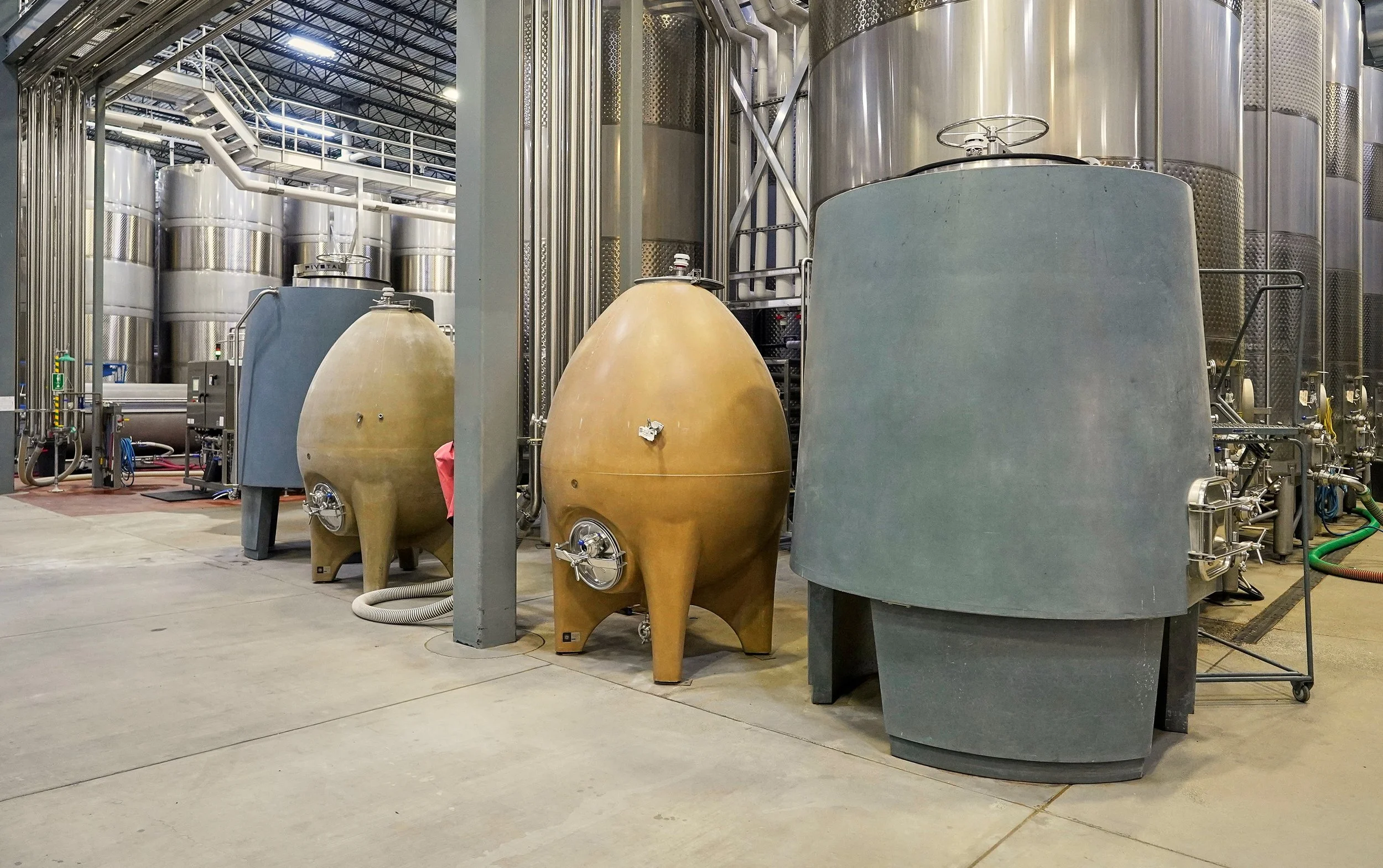Industrial facility with large stainless steel tanks, two tan-colored fermenters or storage tanks, and various pipes and machinery.