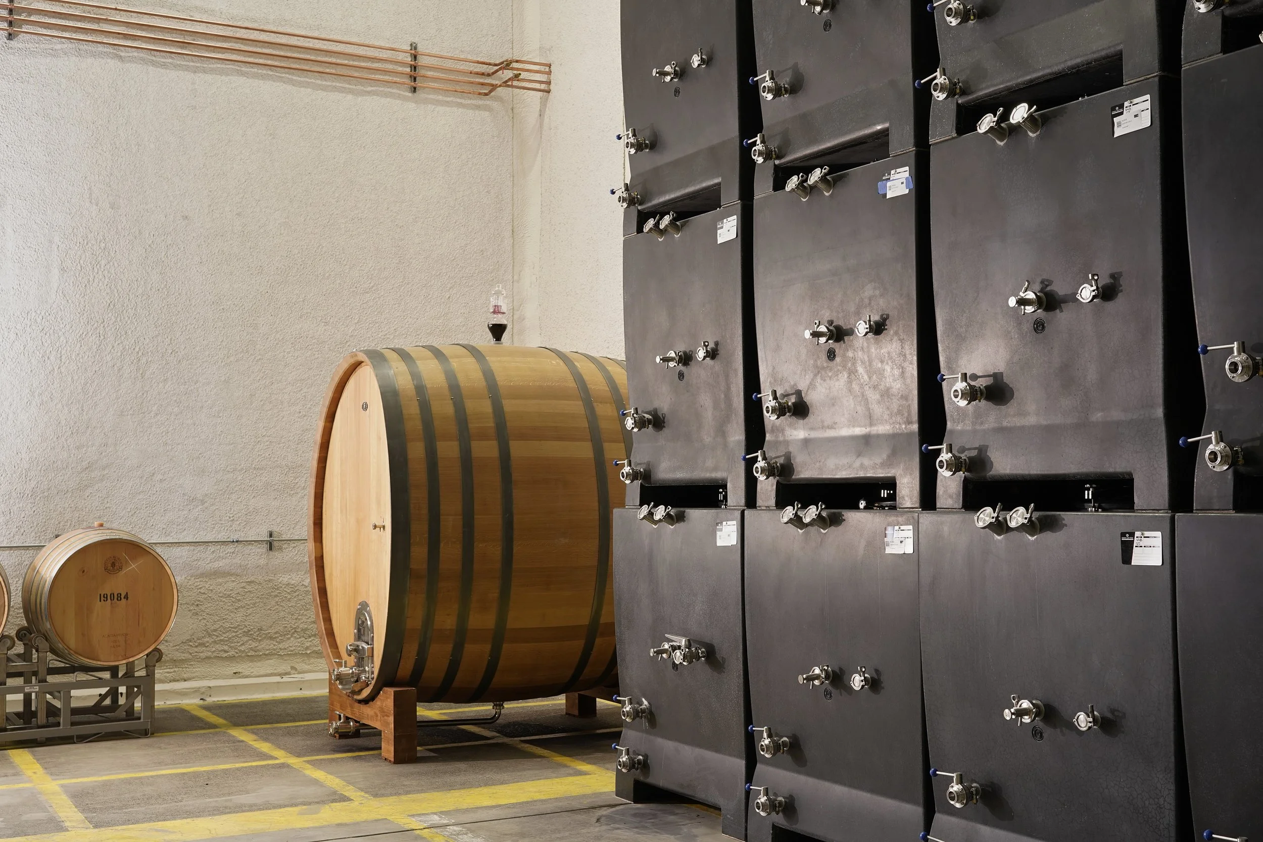 Large black industrial equipment with numerous valves and gauges, two large wooden wine barrels, and some pipes against a beige textured wall in an industrial or storage setting.