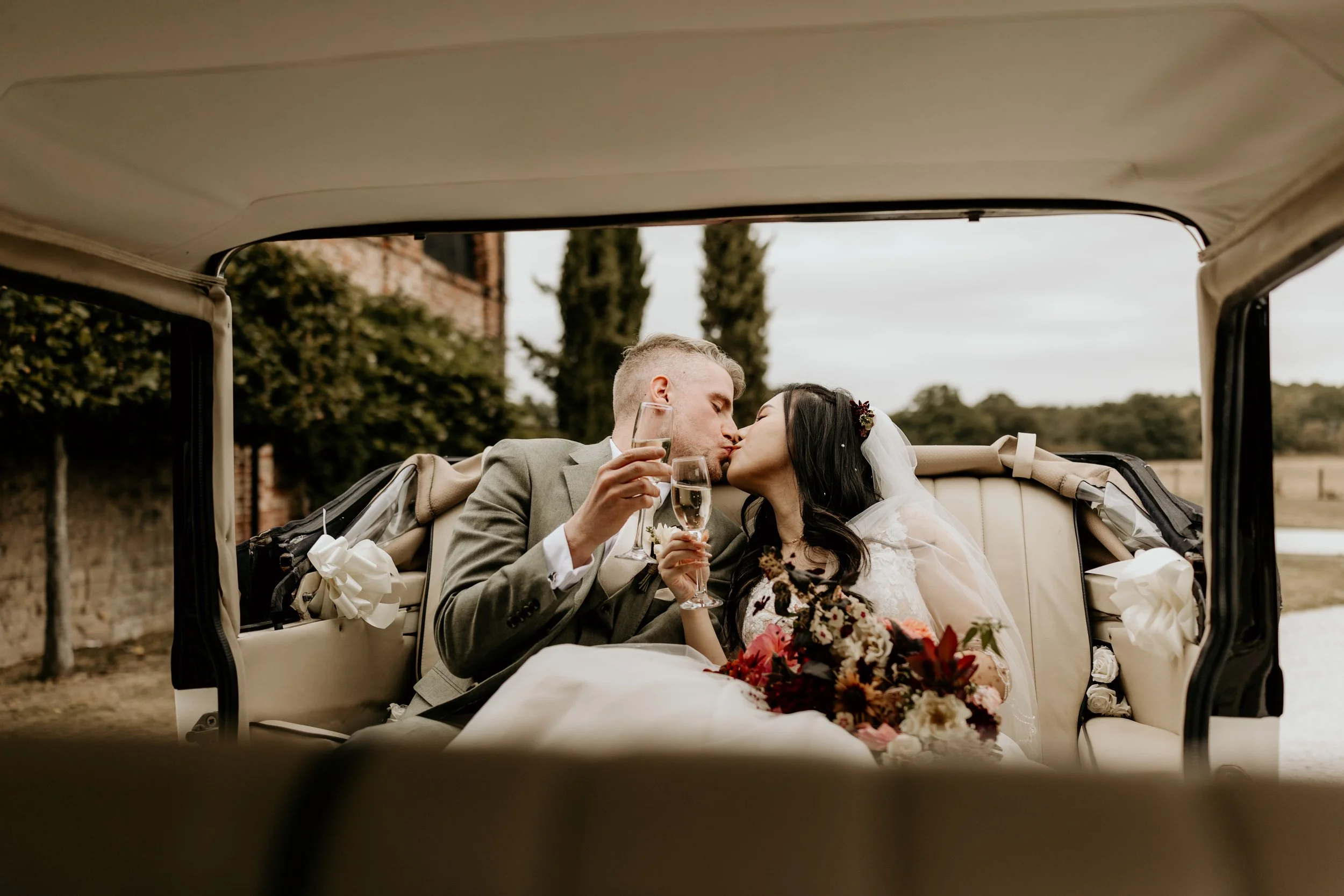 A relaxed wedding day shot of the couple kissing holding a glass of champagne sitting in their wedding car