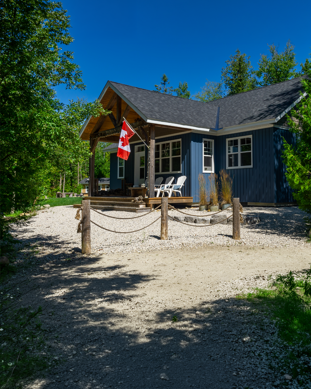 A blue house with a porch, Canadian flag, and outdoor chairs, surrounded by trees under a clear blue sky.