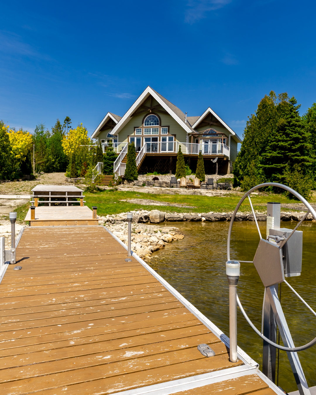 A house by the water with a wooden dock leading to it, surrounded by green trees and shrubs on a clear sunny day.