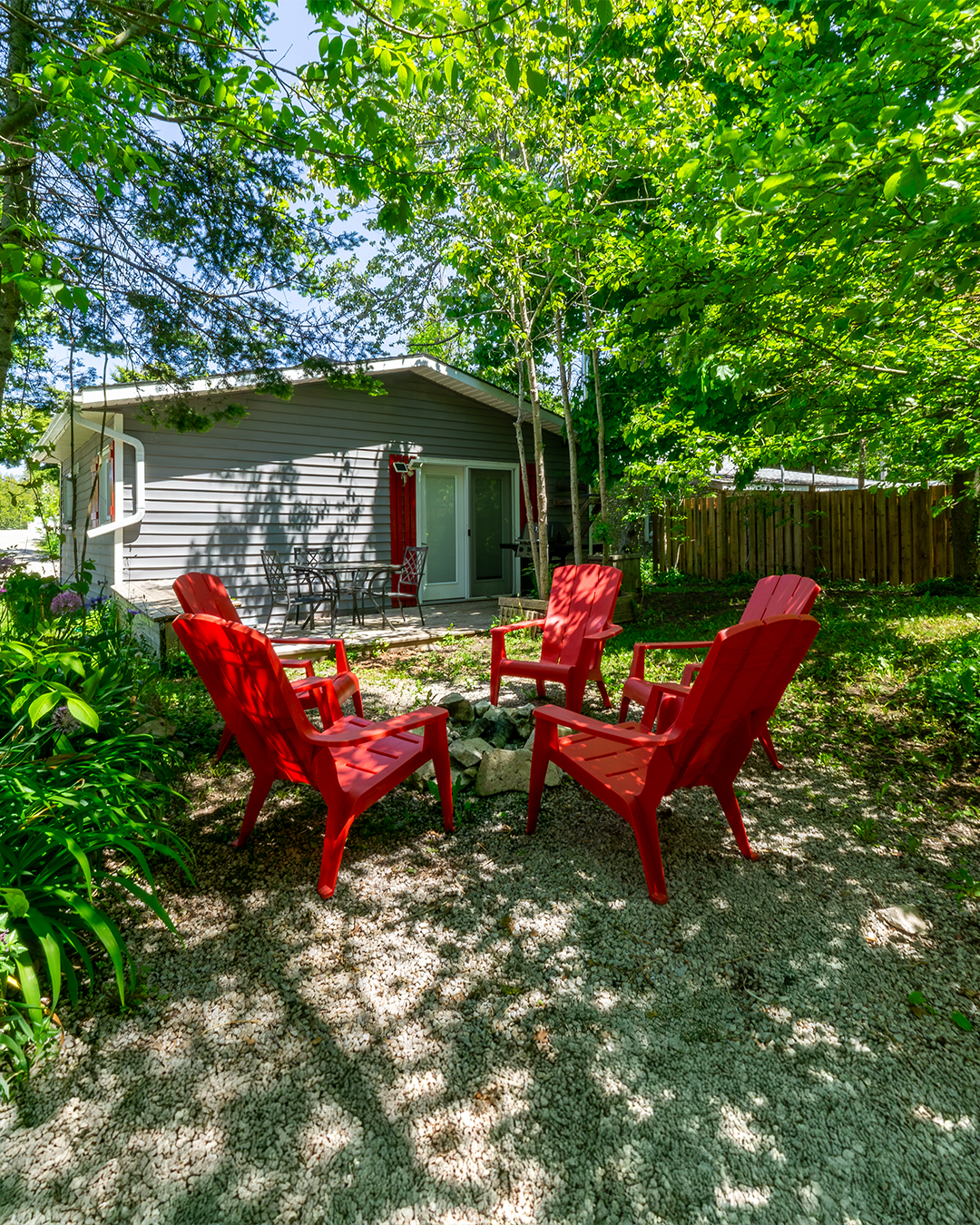 A backyard scene with four red Adirondack chairs arranged in a circle on a gravel area, surrounded by green trees and bushes, with a small house or shed in the background featuring a sliding glass door and outdoor dining table with chairs.