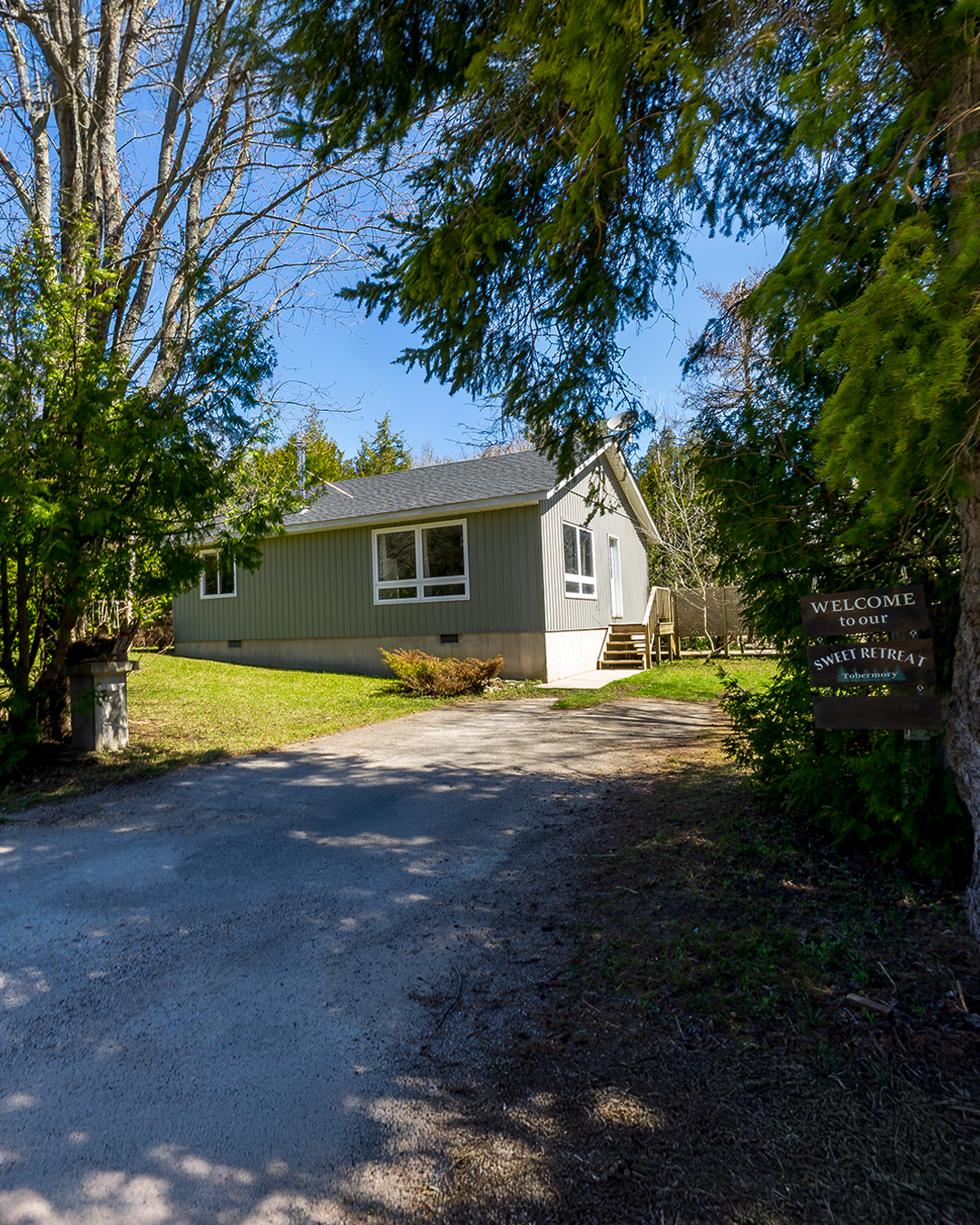 A small grey house with a black roof, surrounded by trees and greenery, with a gravel driveway and a sign that reads 'Welcome to our Sweet Retreat Tobermory'.
