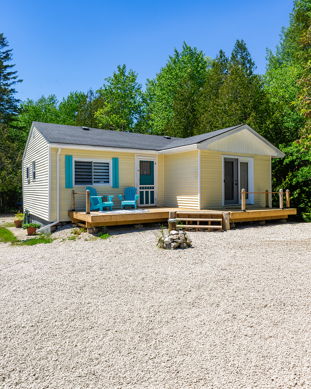 A small, yellow house with a black roof, a wooden deck with two blue Adirondack chairs, and sliding glass doors, surrounded by green trees and a gravel yard.