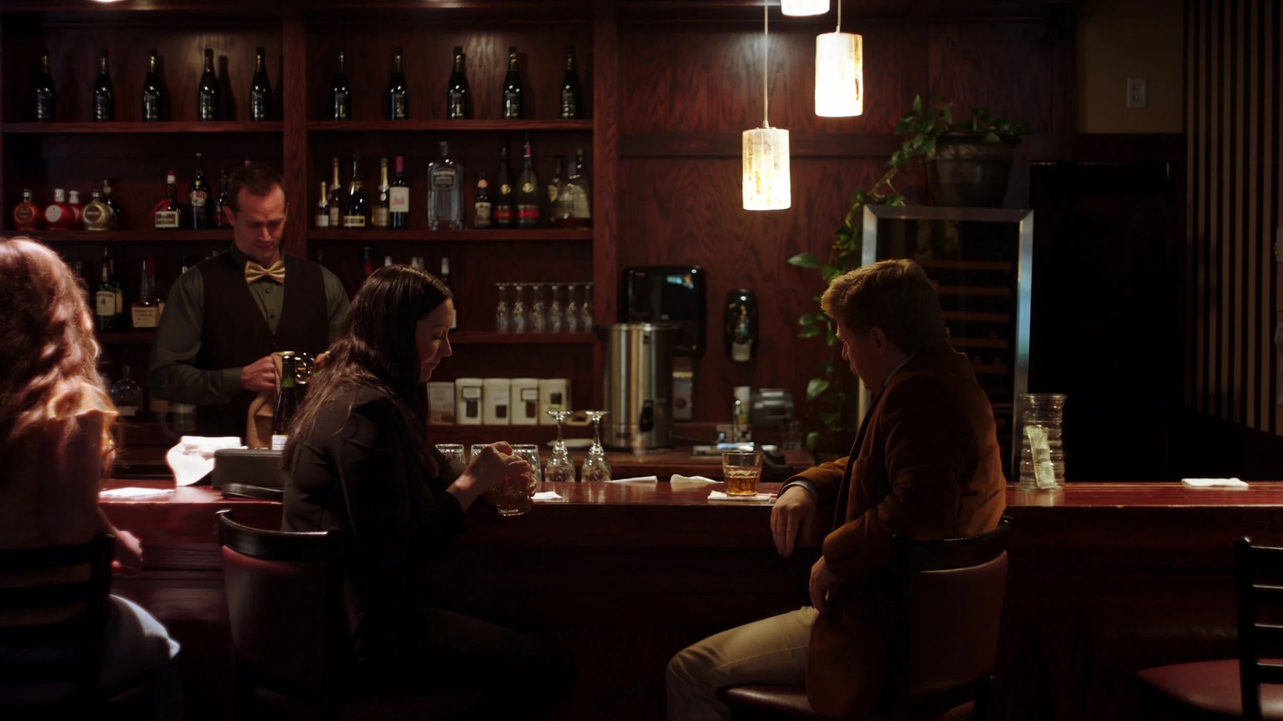 A cozy bar scene with patrons sitting at the counter and a bartender preparing drinks. Dim lighting, wooden shelves with bottles, and hanging pendant lights create a warm atmosphere.