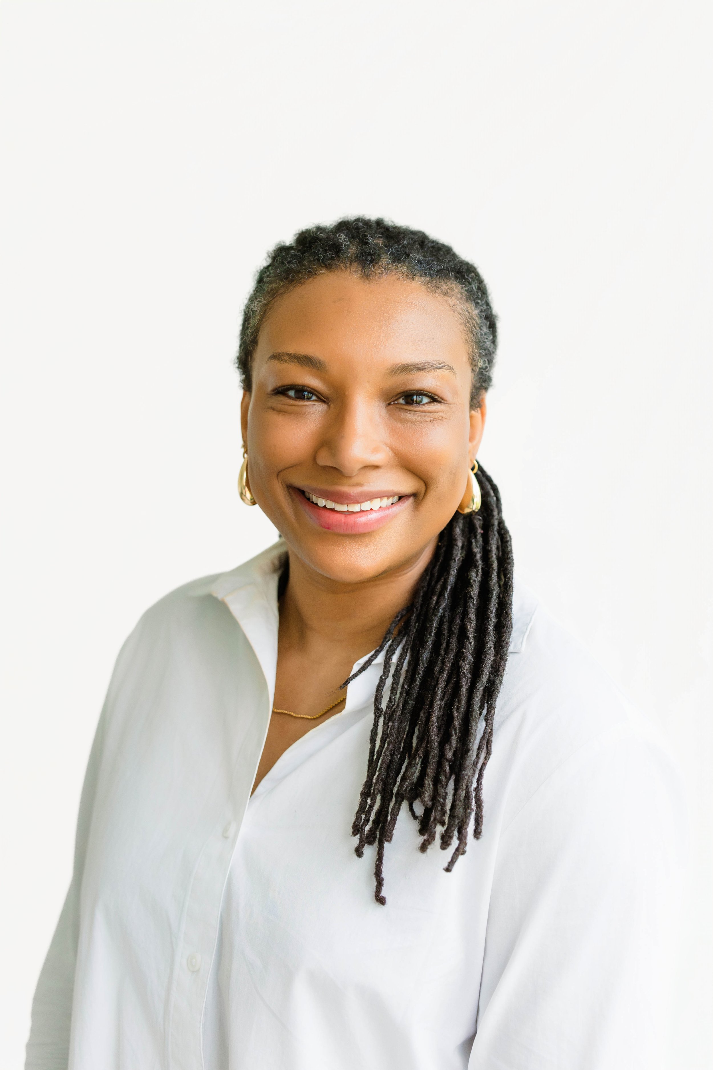 Portrait of a smiling woman with dark hair styled in dreadlocks, wearing large gold hoop earrings, a dark blazer, and a white shirt, against a plain white background.