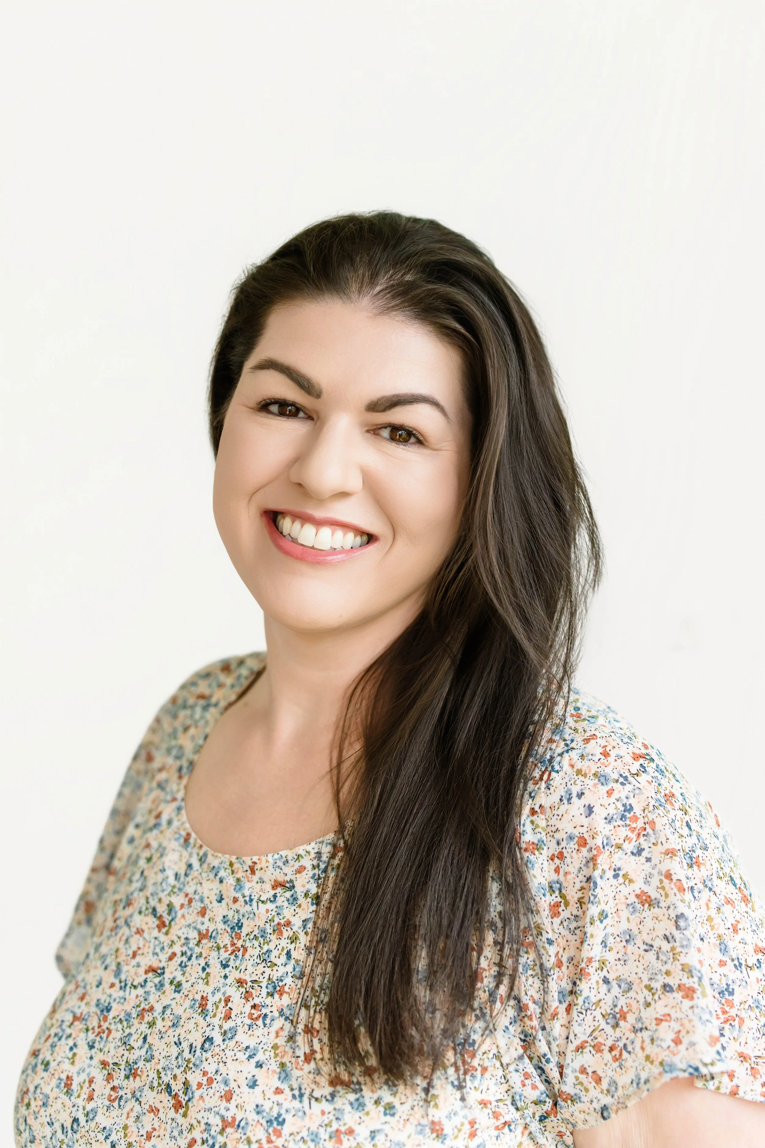 Close-up of a woman with long, wavy brown hair, smiling and showing teeth, wearing a white top with black dots, against a white background.