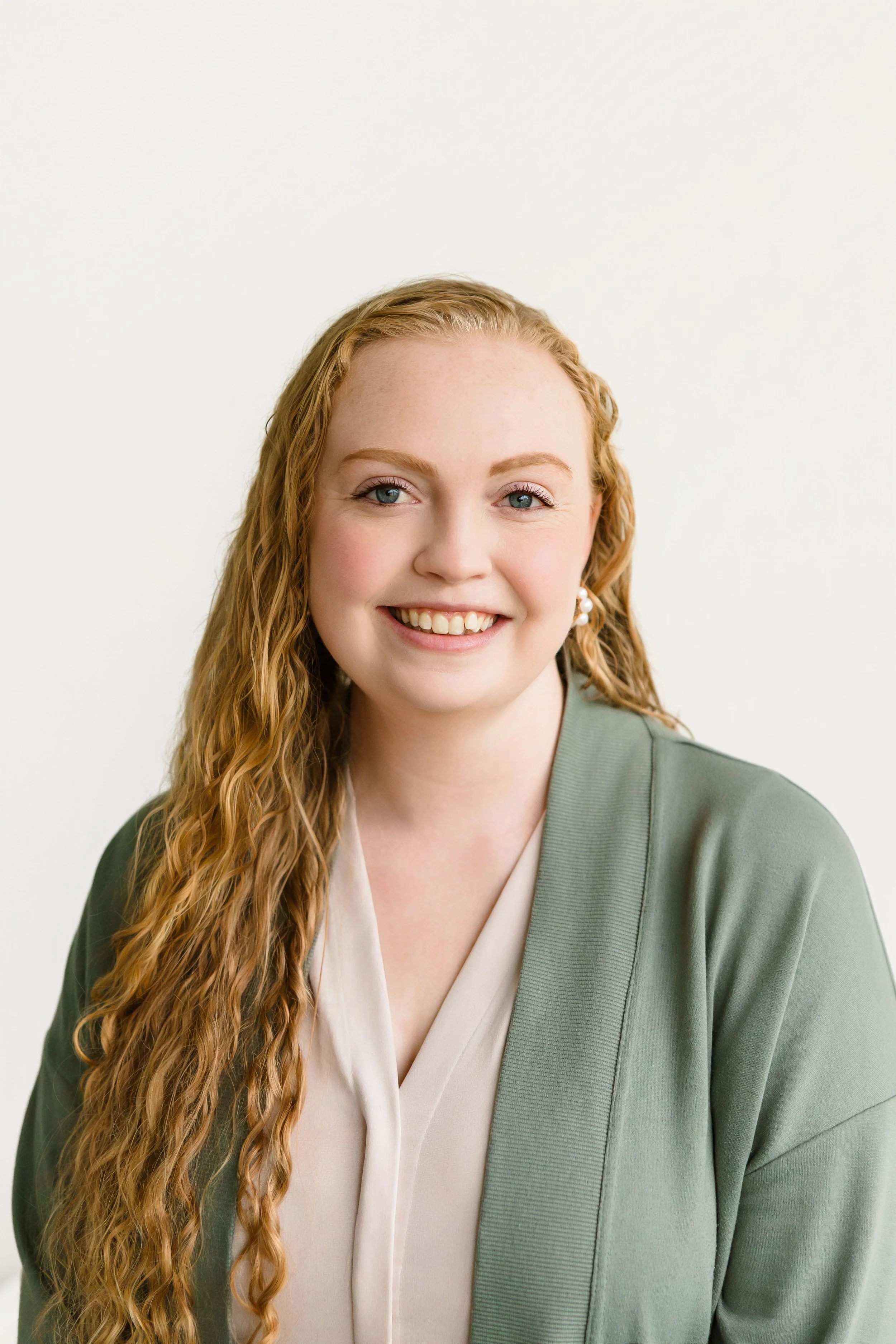 Portrait of a young woman with curly red hair, smiling, wearing a dark green top and a silver necklace, against a plain white background.