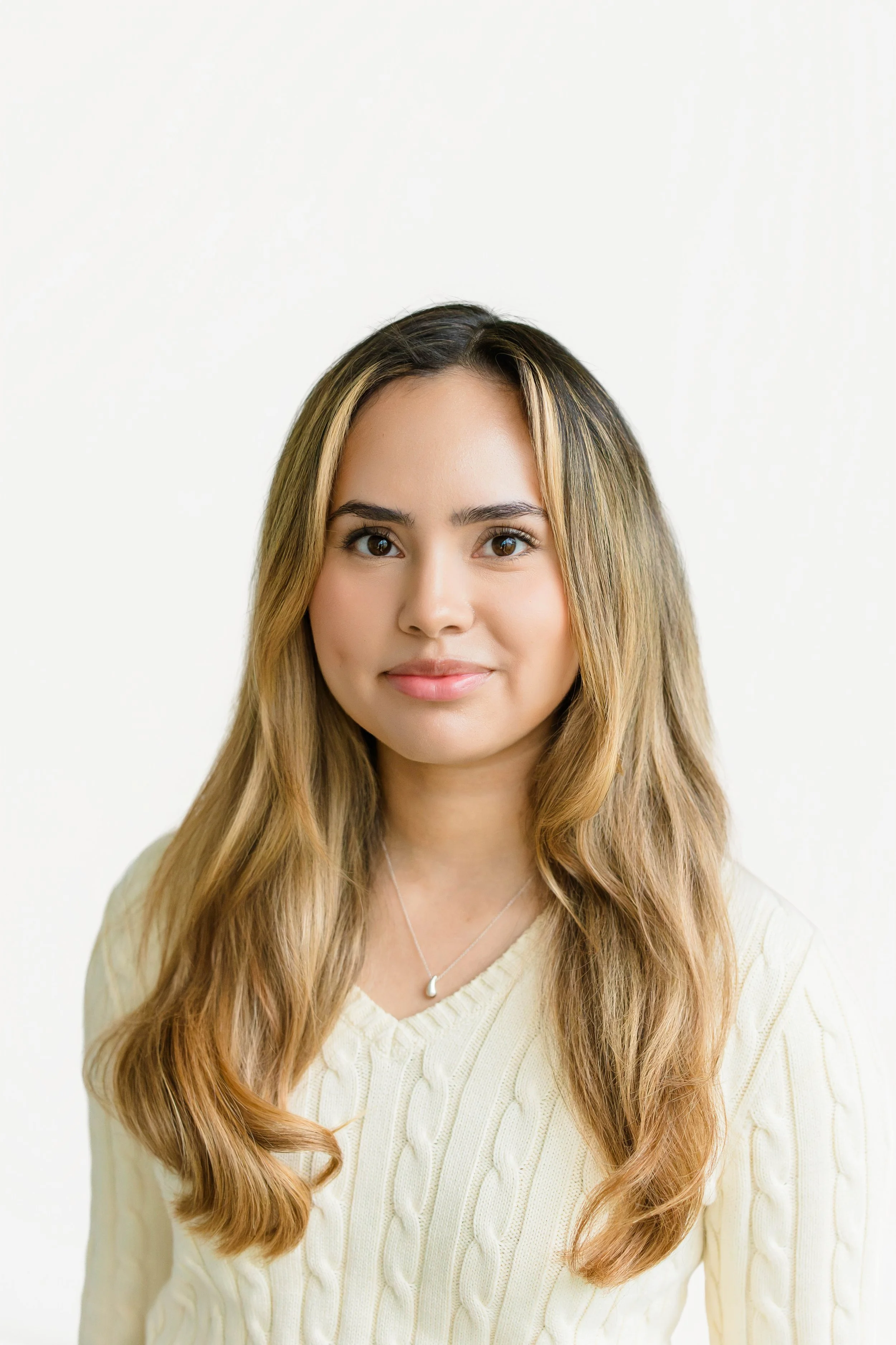 Portrait of a young woman with long wavy hair, smiling, wearing a white off-the-shoulder top and layered necklaces, against a plain white background.